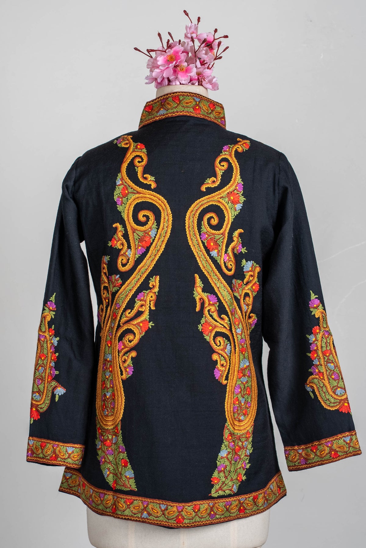 Full back view of the black short jacket on a mannequin, with long mirrored golden-orange Aari scrolls and colourful embroidered borders