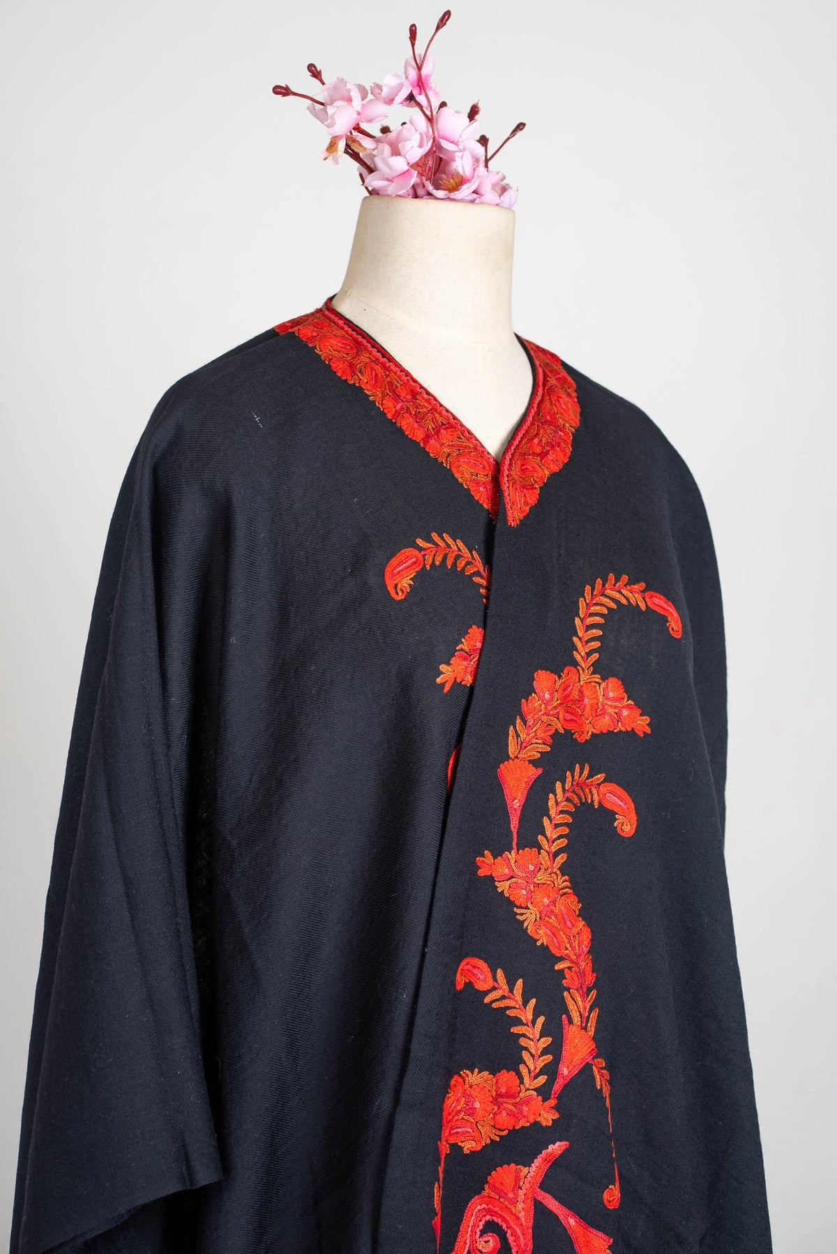 Side view of the black cape across a mannequin, showing its loose drape and red-orange embroidered neckline and front