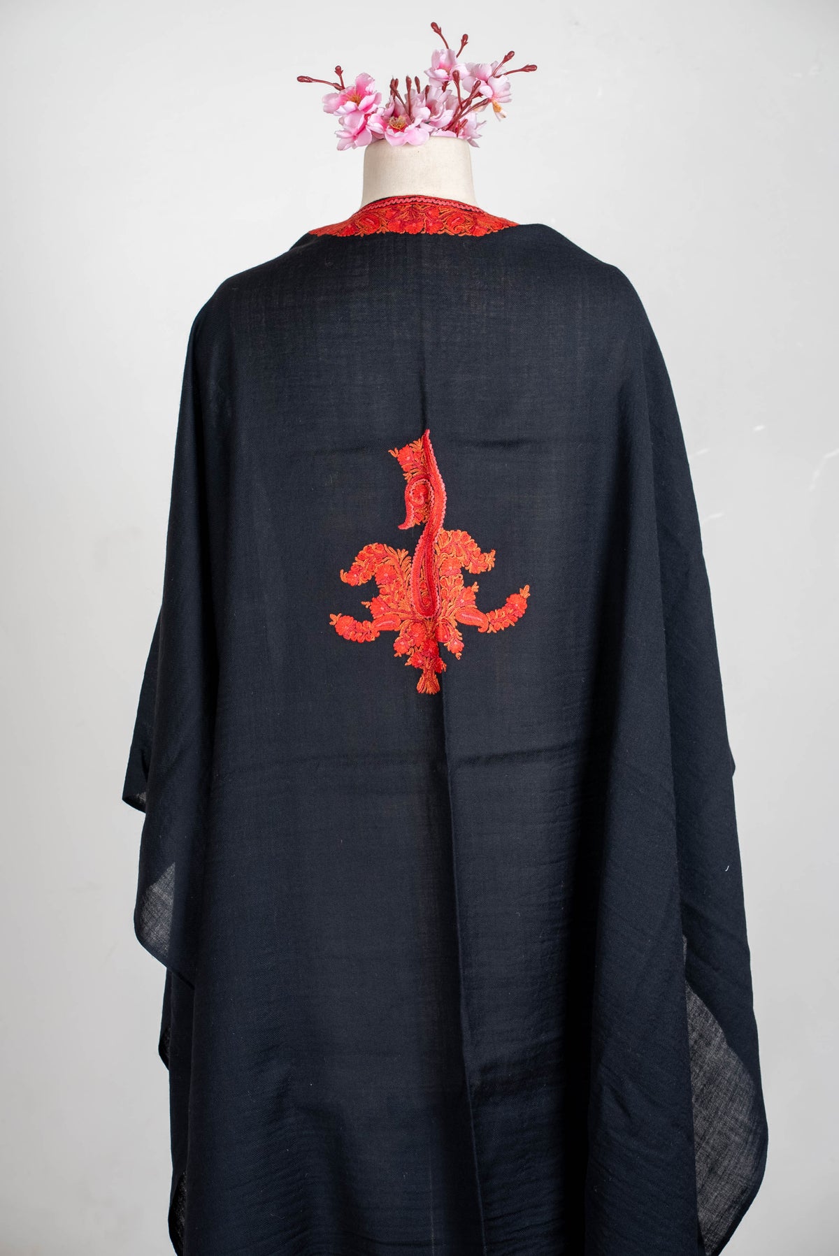Back view of the black cape on a mannequin, with a single red-orange embroidered motif between the shoulders