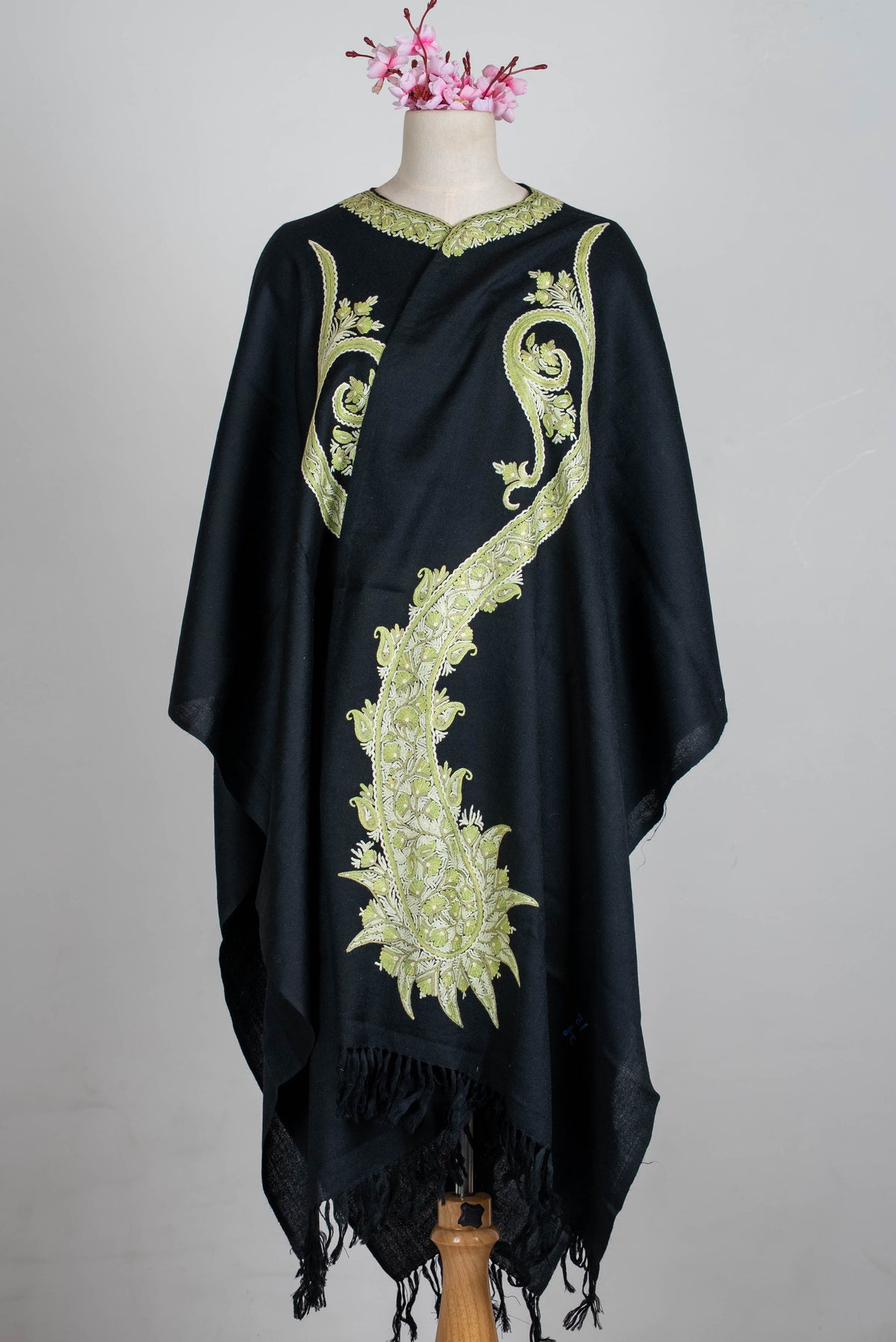 Full front view of the black cape wrapped across a mannequin, with elongated sage-green paisley embroidery and black fringe