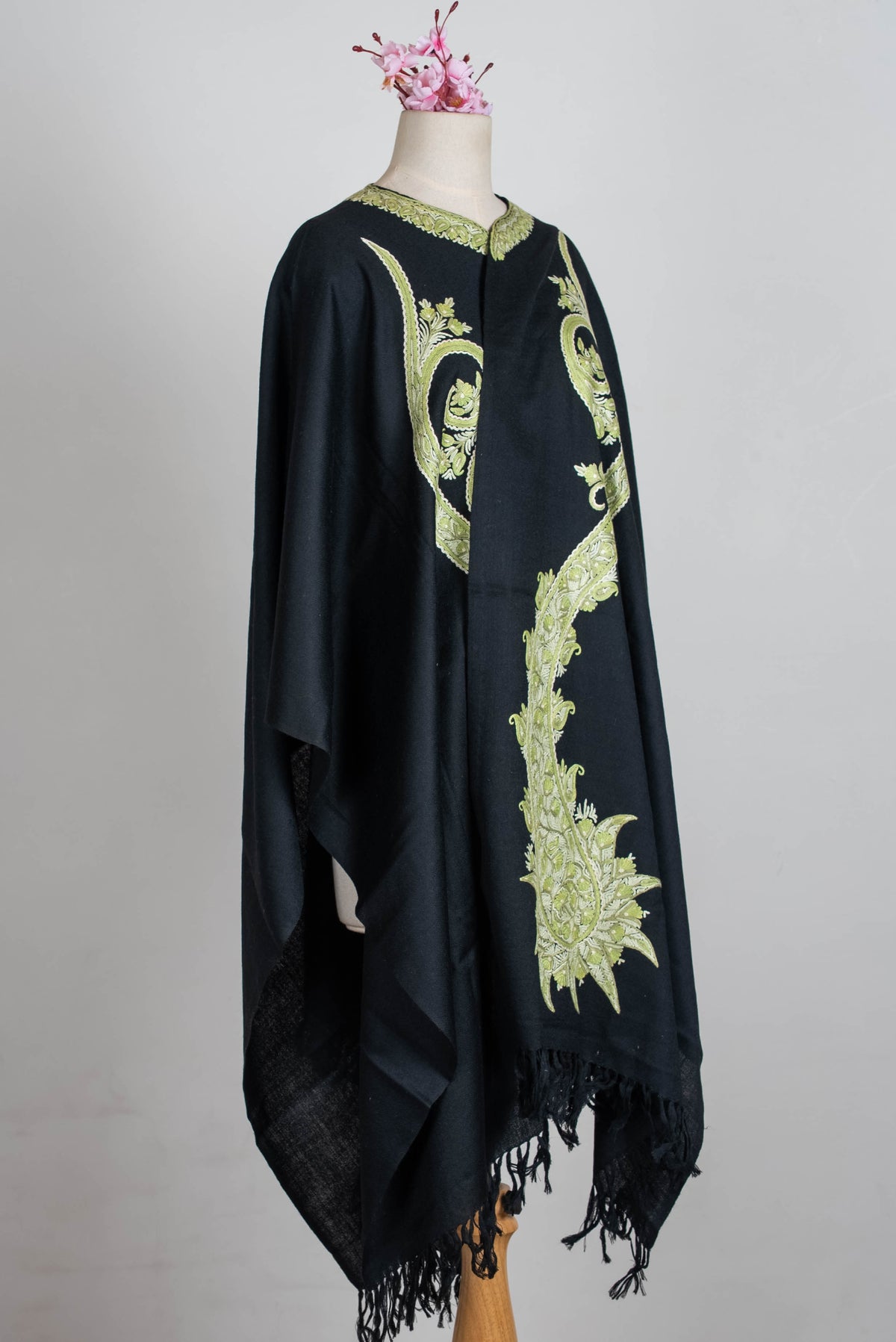 Angled full view of the black cape on a mannequin, showing long sage-green embroidered paisleys and black fringed ends