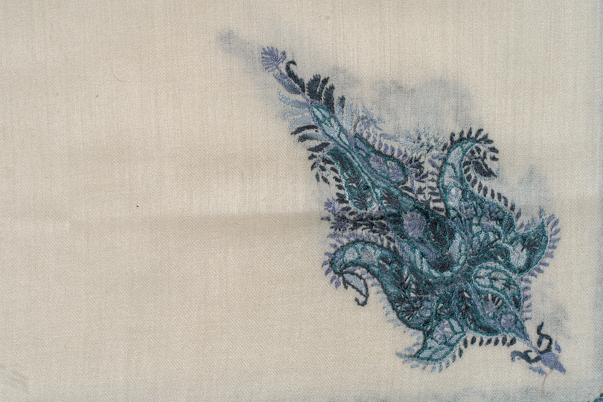 Close-up of an ornate teal paisley motif with curling leaves and fine floral stitching on ivory fabric