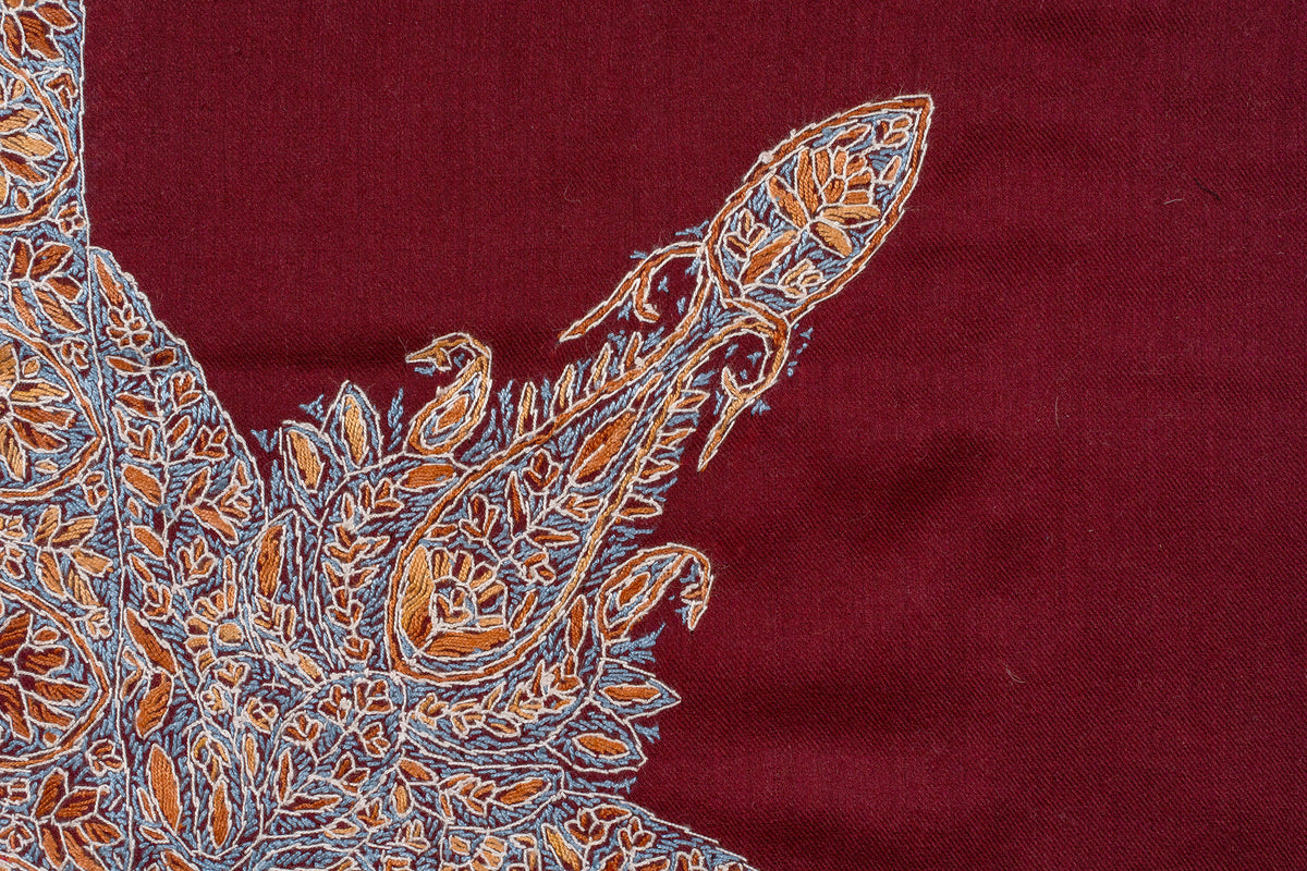 Close-up of a pointed orange-and-ivory Sozni paisley surrounded by fine pale blue stitching on crimson fabric