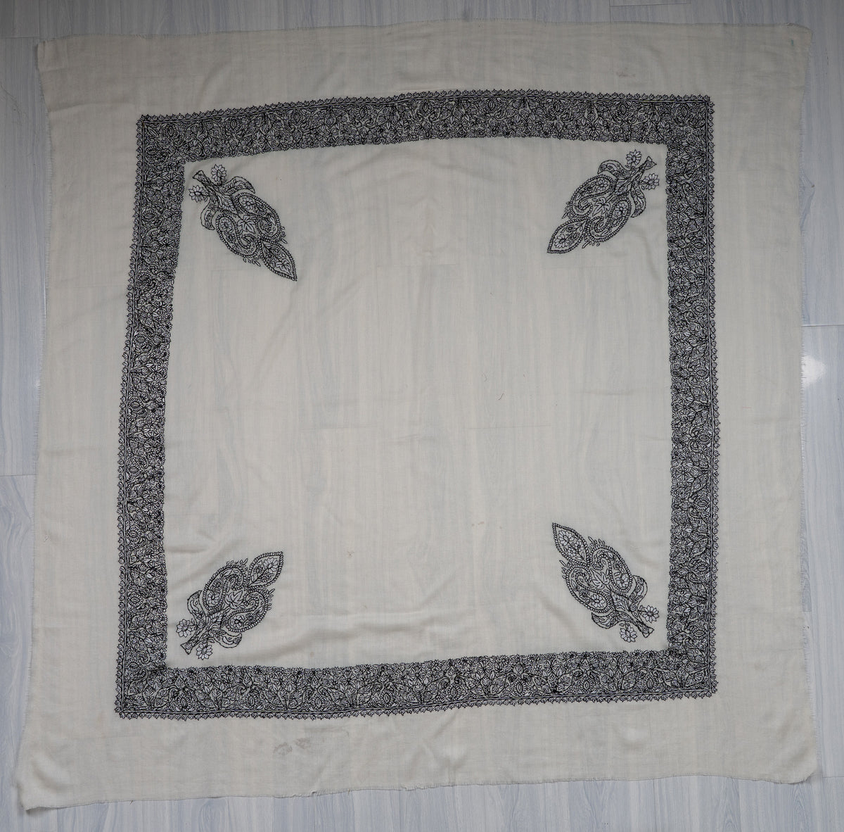 Square ivory Merino wool rumal laid flat with black Sozni borders, four corner motifs and a plain centre