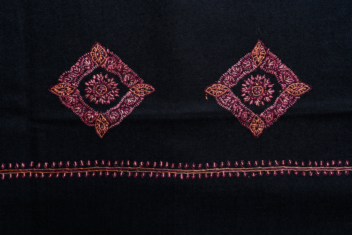 Flat detail of two red diamond-shaped floral medallions above the fine embroidered border on the black shawl.