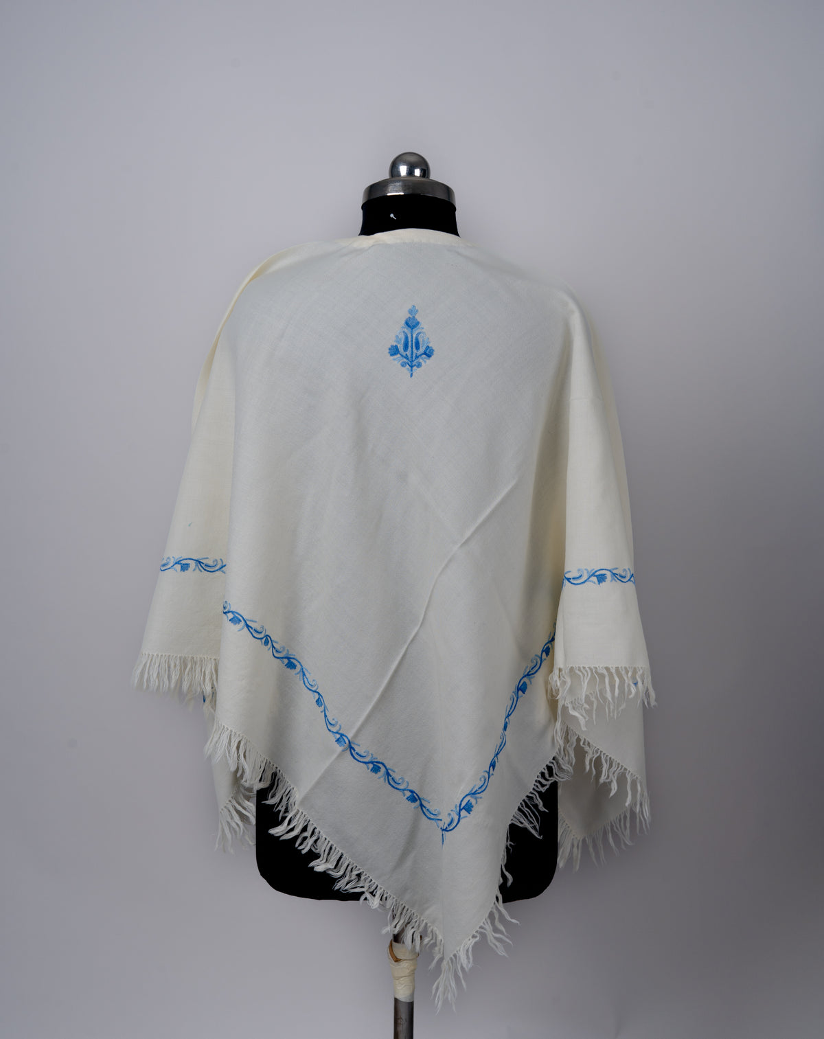 Back view of the ivory poncho with a small blue motif near the neck and a narrow embroidered border above the pointed fringed hem.