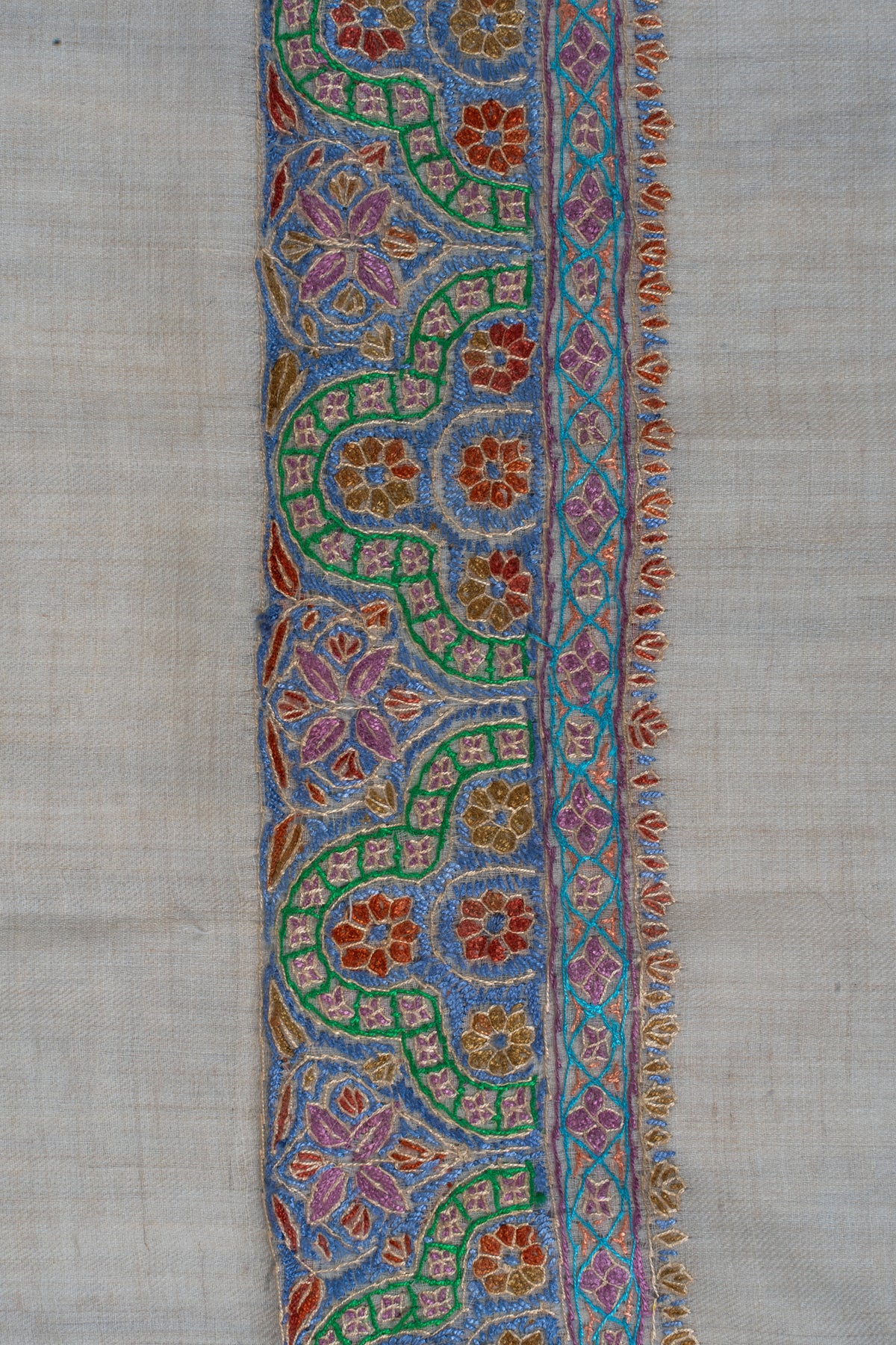 Close-up of a Sozni border with blue panels, green curves, pink flowers and orange accents on cream cashmere.