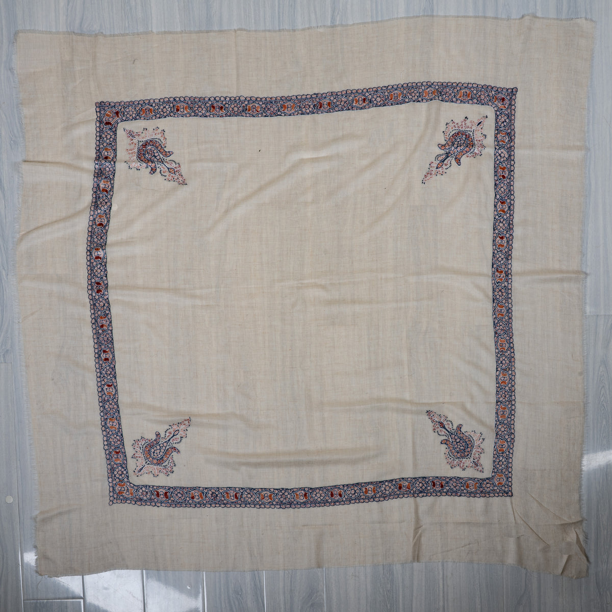 Ivory square cashmere rumal laid flat, with an indigo patterned border and four coral and rust floral corner motifs