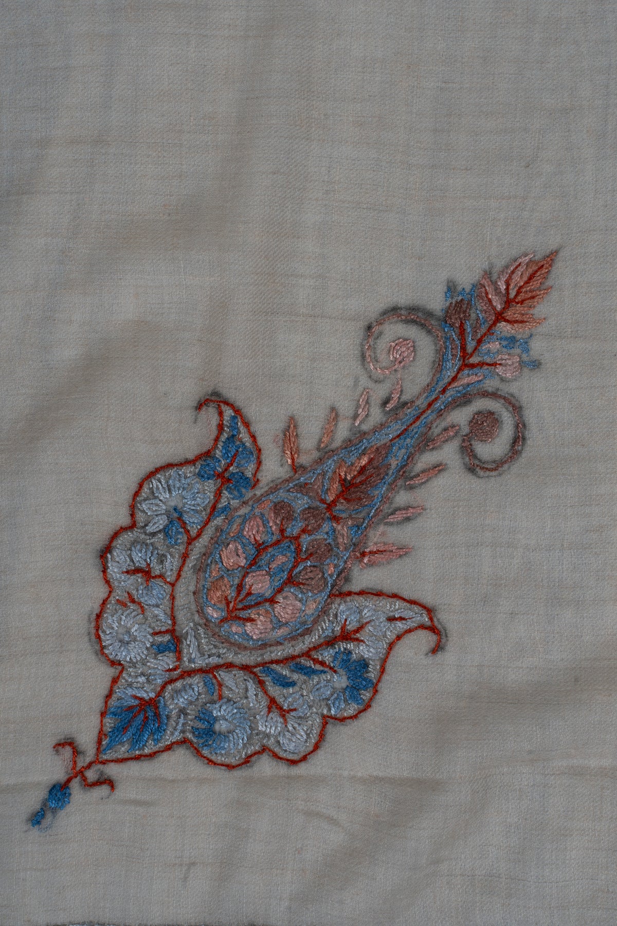 Close-up of a small coral and powder-blue Sozni paisley with curling tips and fine leaf details on beige fabric