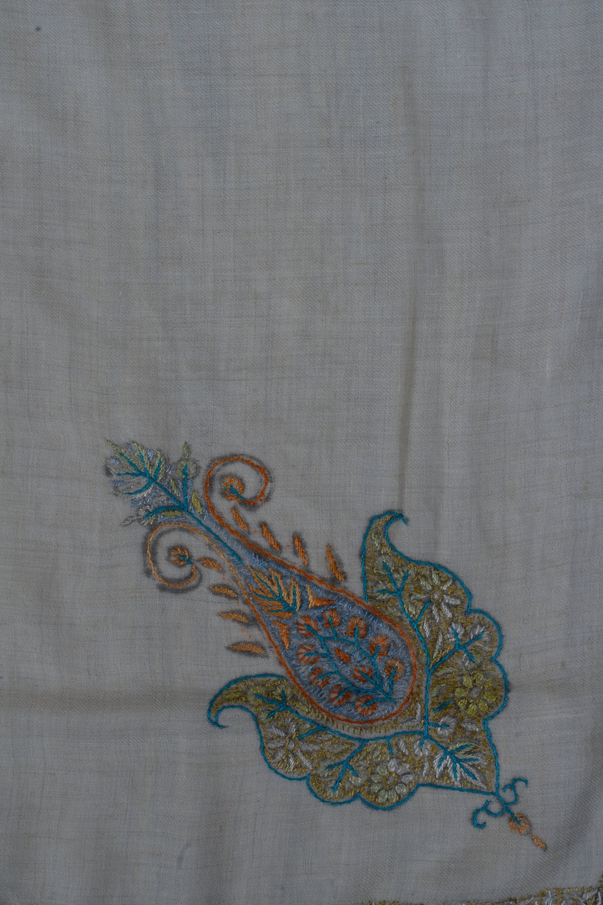 Close-up of an amber and blue Sozni paisley with a curled tip and muted green floral details on pale cashmere.
