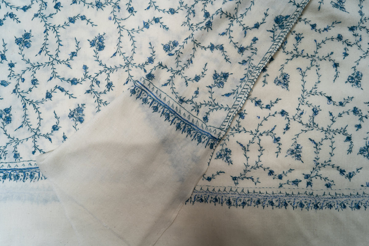 Folded corner of the ivory Sozni stole, showing blue floral embroidery on both sides and a narrow leaf-edged border