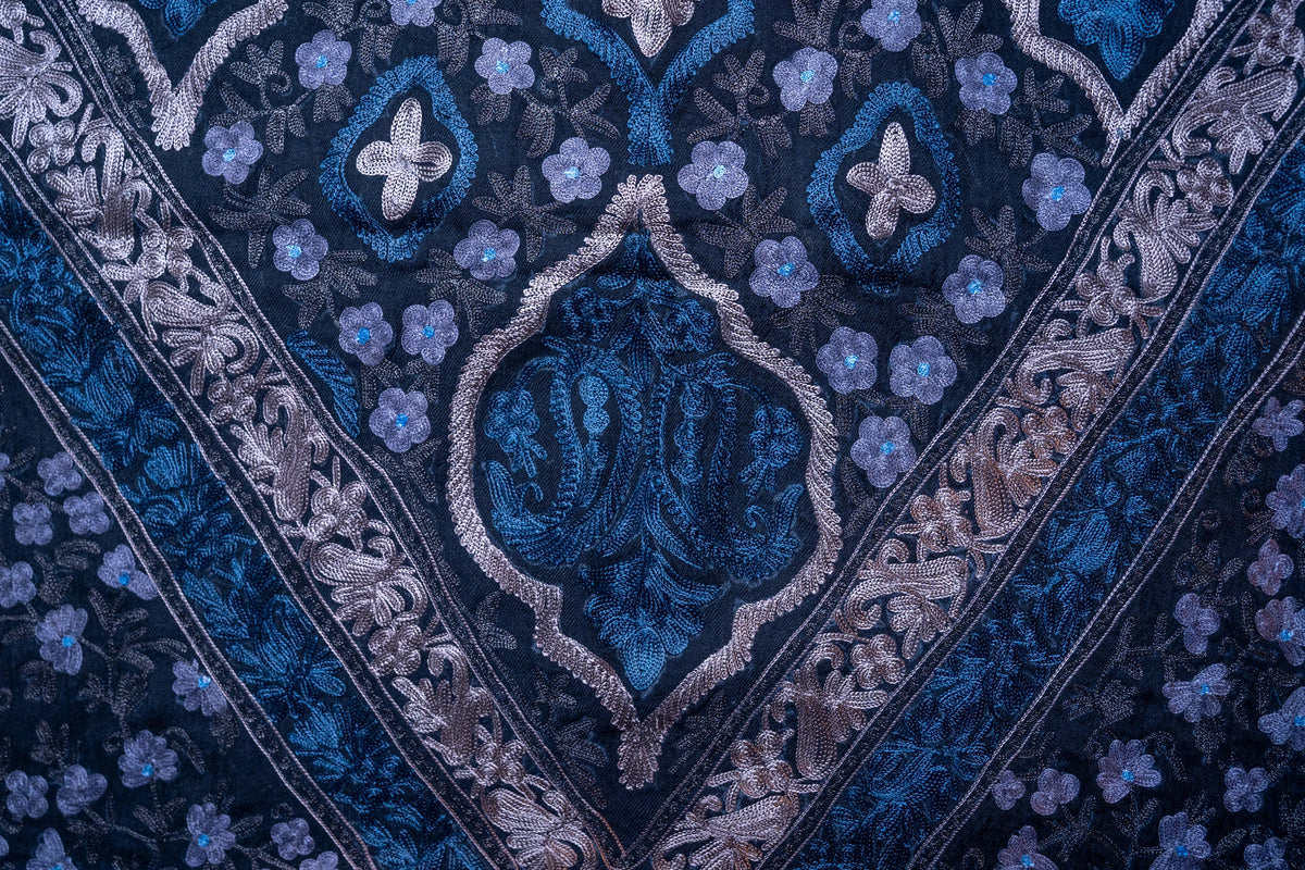 Close-up of a blue floral medallion outlined in silver-toned stitching beside small blue flowers on black fabric