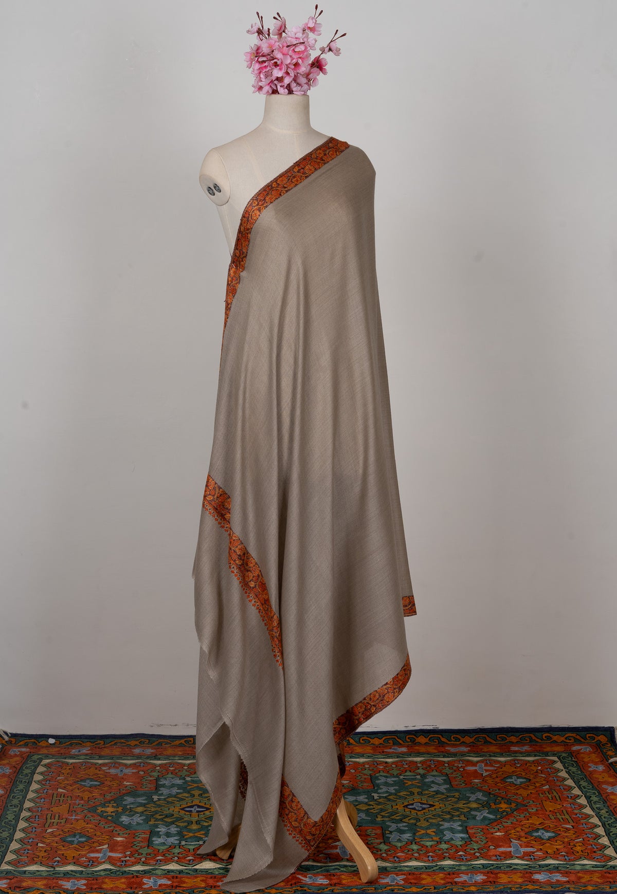 Full-length grey-beige shawl on a mannequin with narrow orange-red embroidered borders around the plain fabric