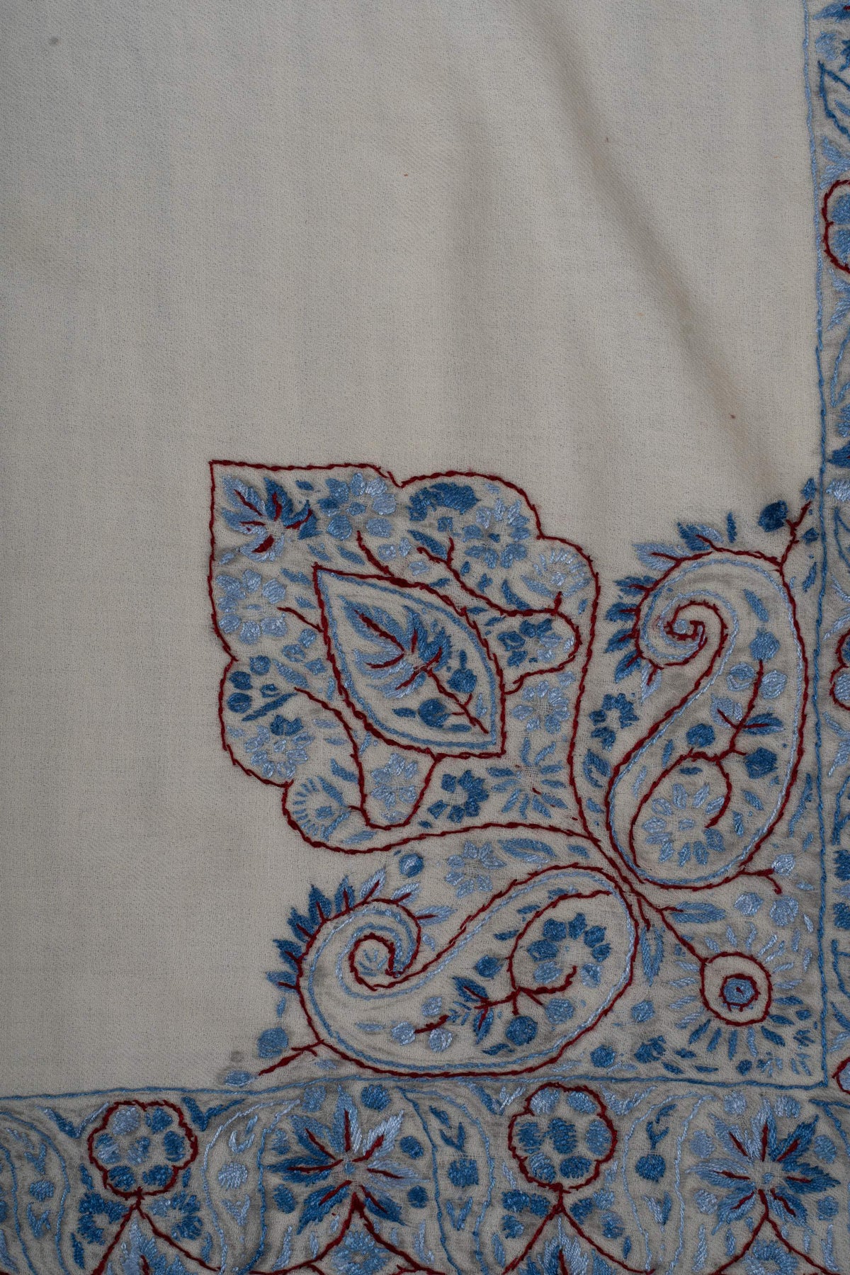 Close-up of a Sozni corner motif with blue flowers, curled paisleys and a burgundy-outlined leaf on ivory Merino wool.