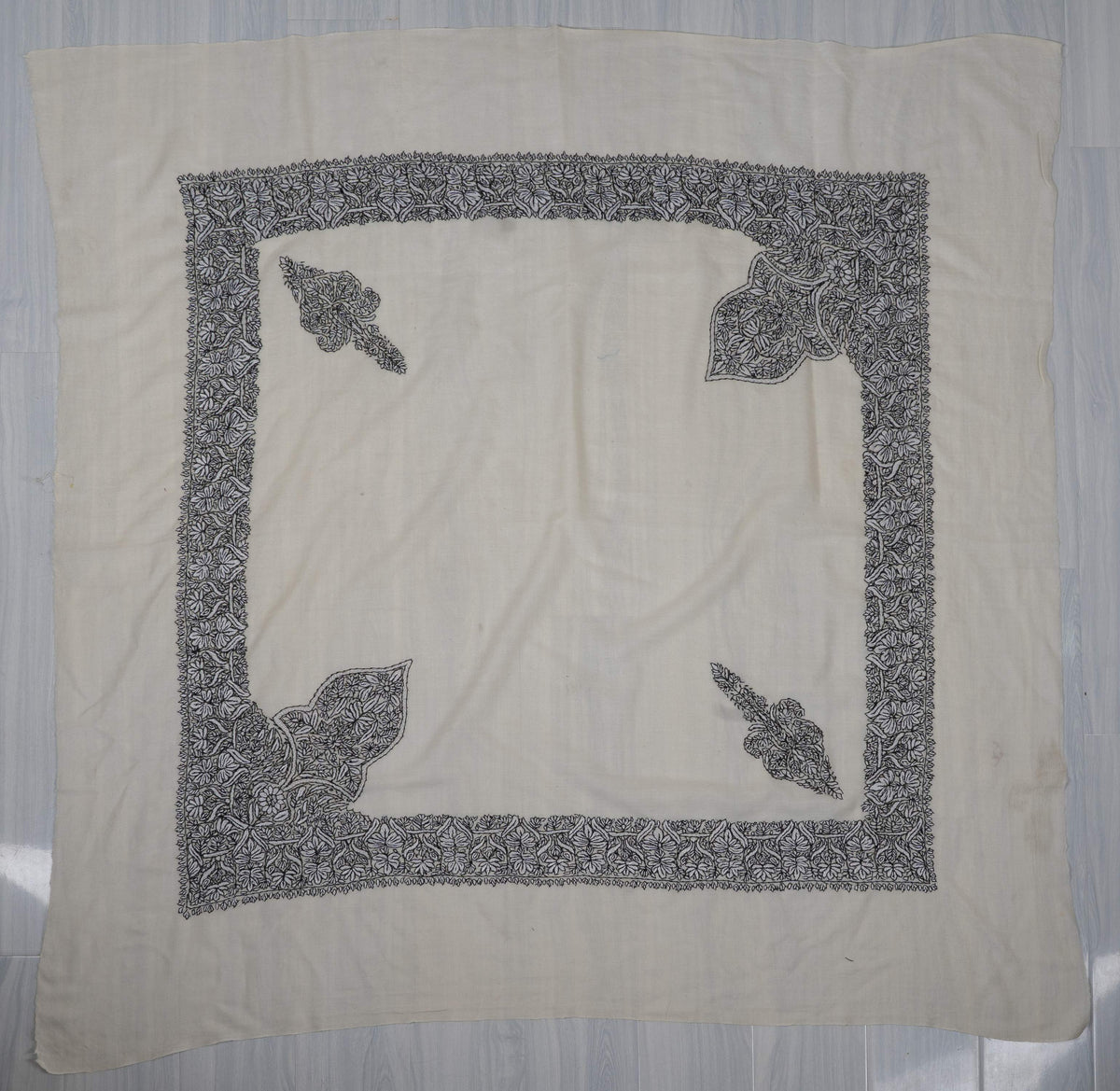 Ivory square Merino wool rumal laid flat, with a broad black floral border and four differently shaped corner motifs