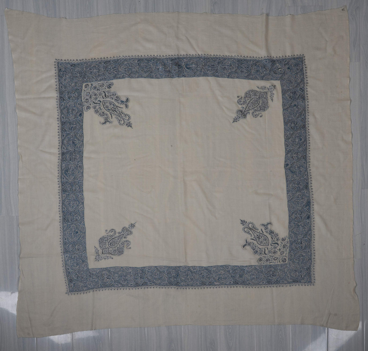 Ivory square rumal spread flat with an indigo floral border, four paisley corner motifs and a plain centre.
