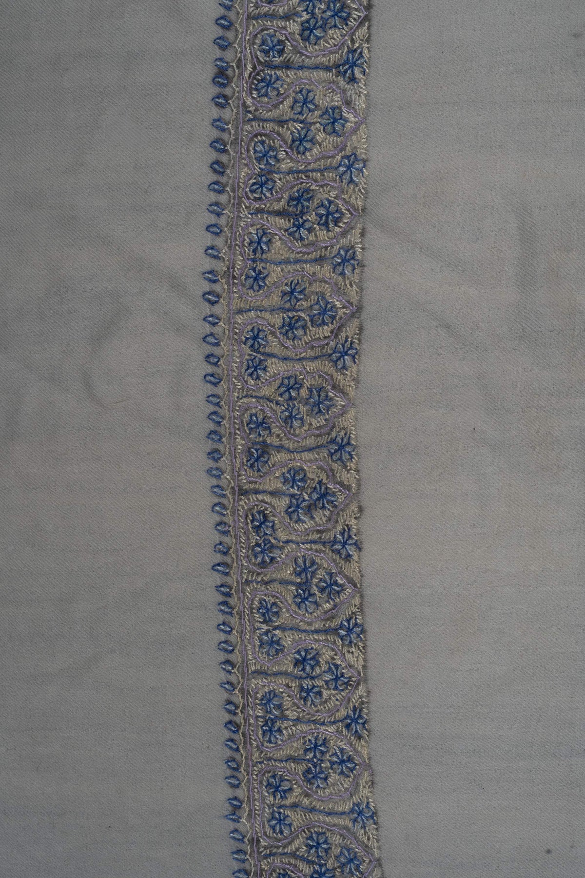 Close-up of blue Sozni flowers and pale curling stems with tiny leaf edging on an ivory Merino wool rumal border.