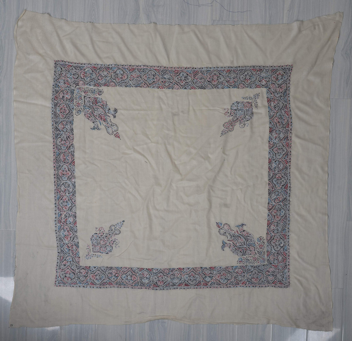 Ivory Merino rumal laid flat, with blue-pink floral borders and two pairs of different corner motifs
