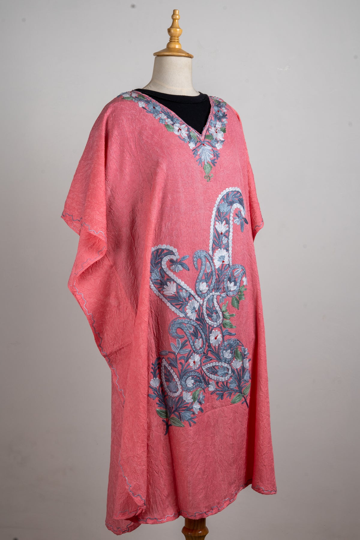 Angled full view of a coral-pink kaftan on a mannequin, with loose sleeves and large blue-and-white paisley embroidery