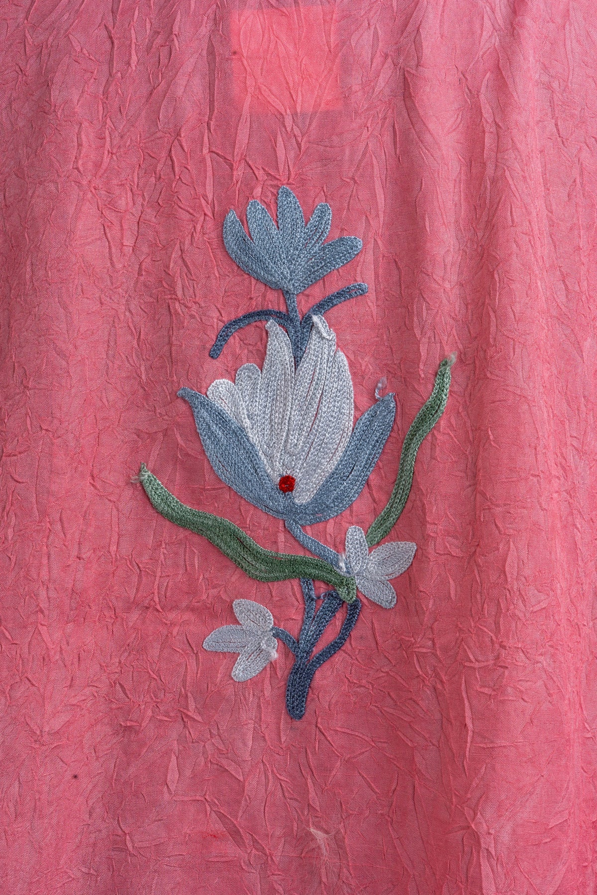 Close-up of a small blue-and-white Aari flower motif with green leaves on the coral-pink kaftan’s back