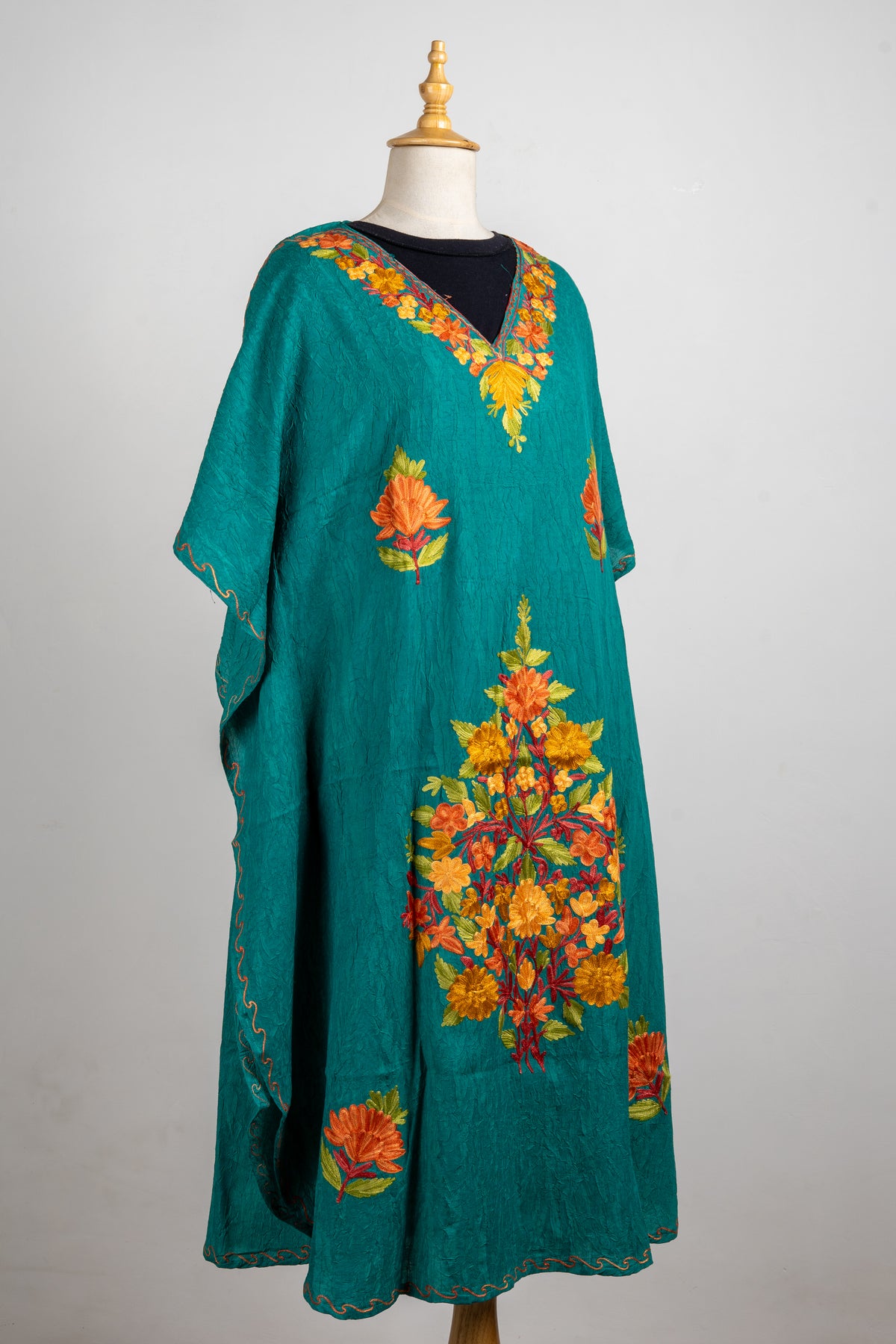 Angled full view of the teal kaftan with loose sleeves, scattered flowers and a large orange and yellow central bouquet.