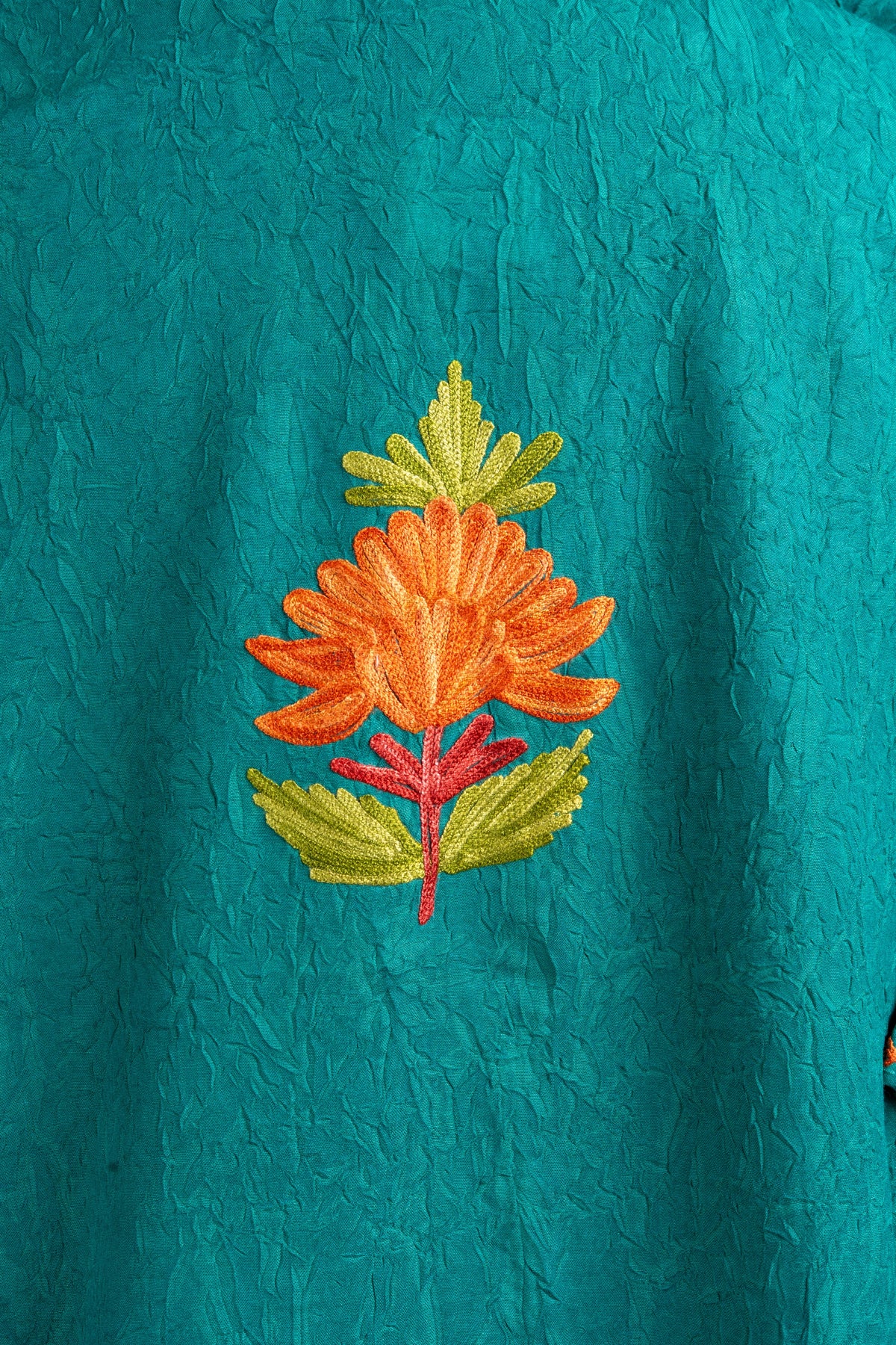 Detail of a single orange embroidered flower with green leaves and a red stem on textured teal fabric.