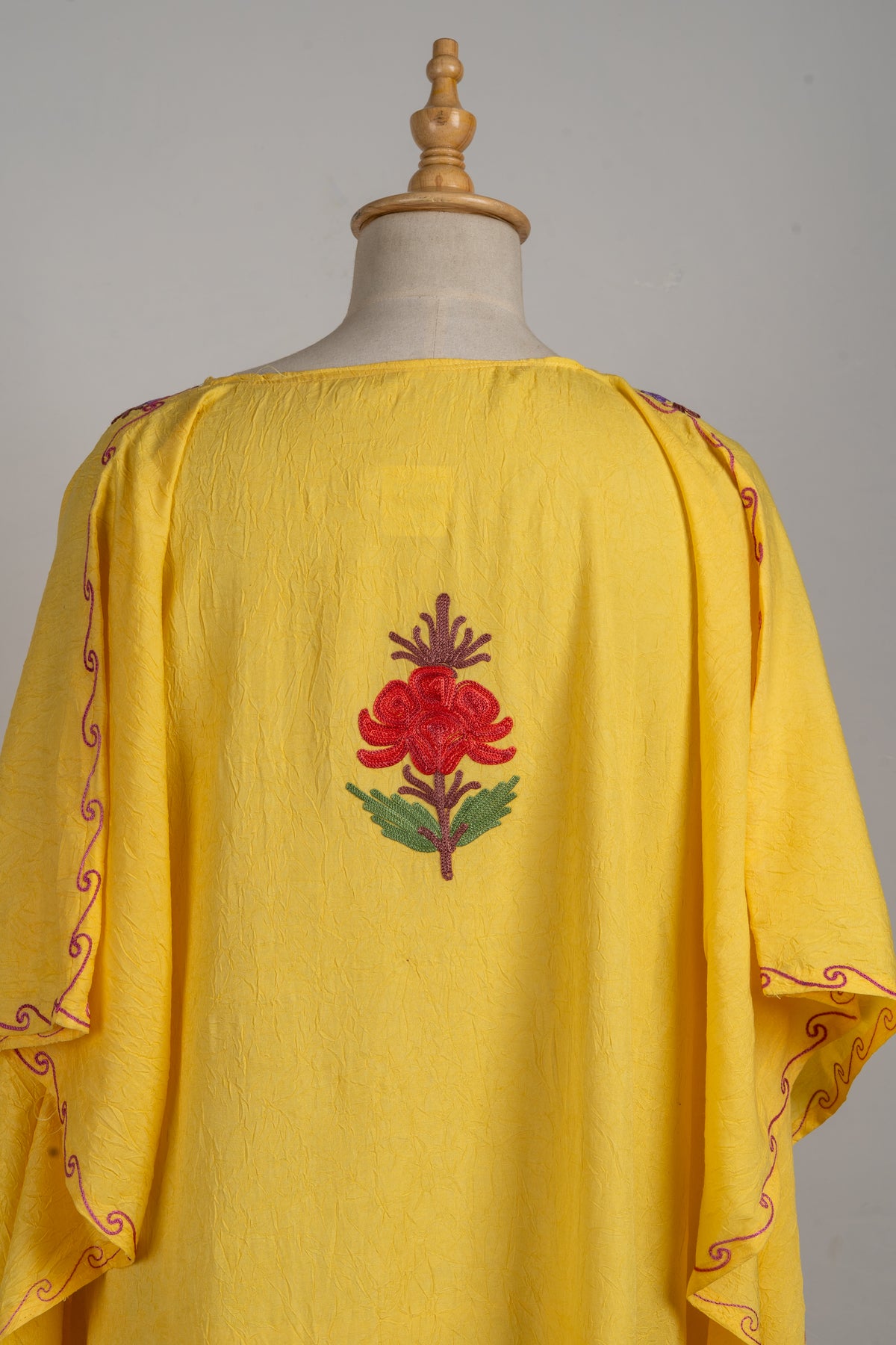 Upper back of a yellow kaftan with one red Aari flower, green leaves and a purple stem between the draped sleeves