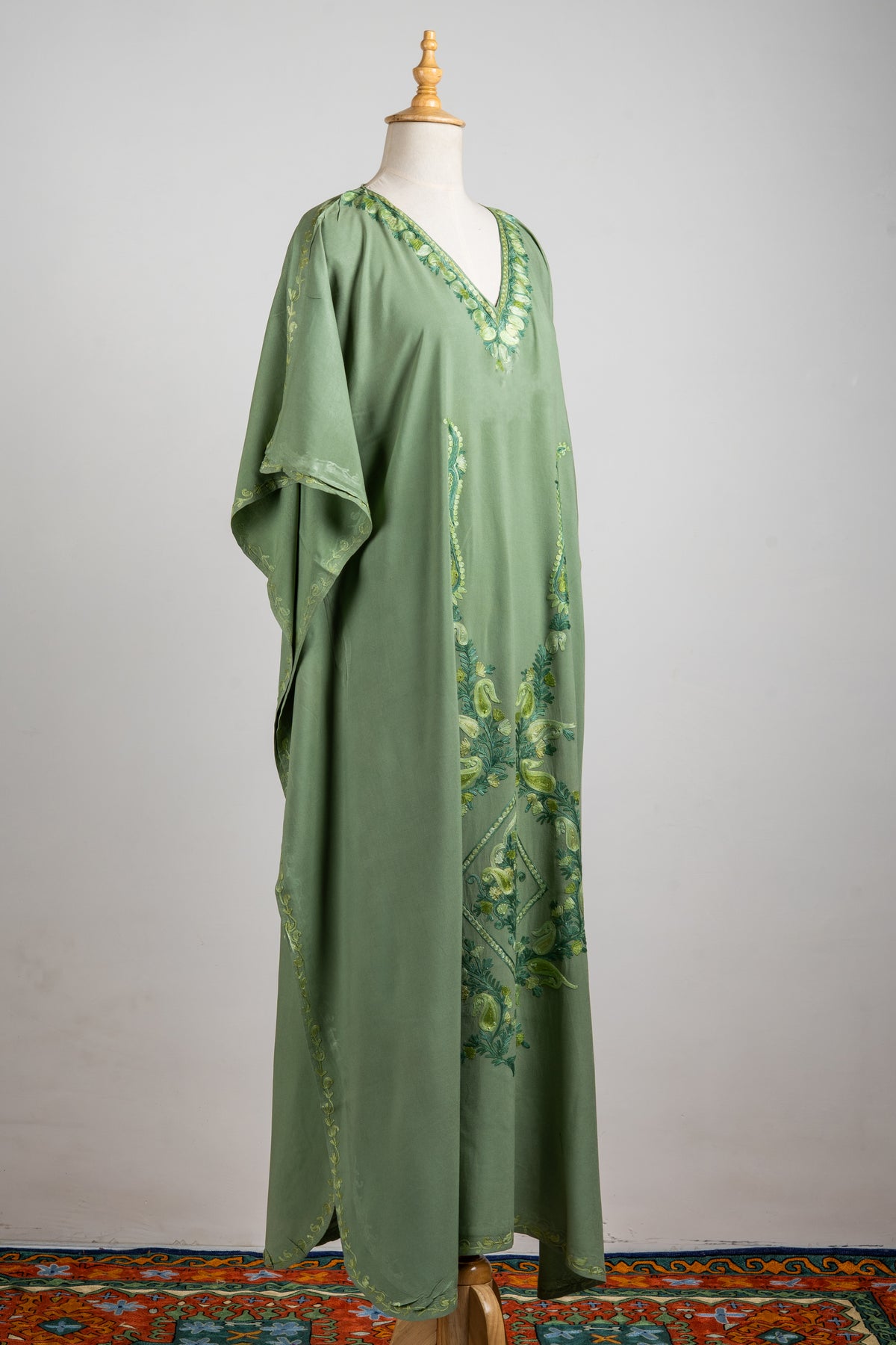 Angled full view of the olive-green kaftan showing loose sleeves, a leafy V-neck border and a long botanical design down the front.