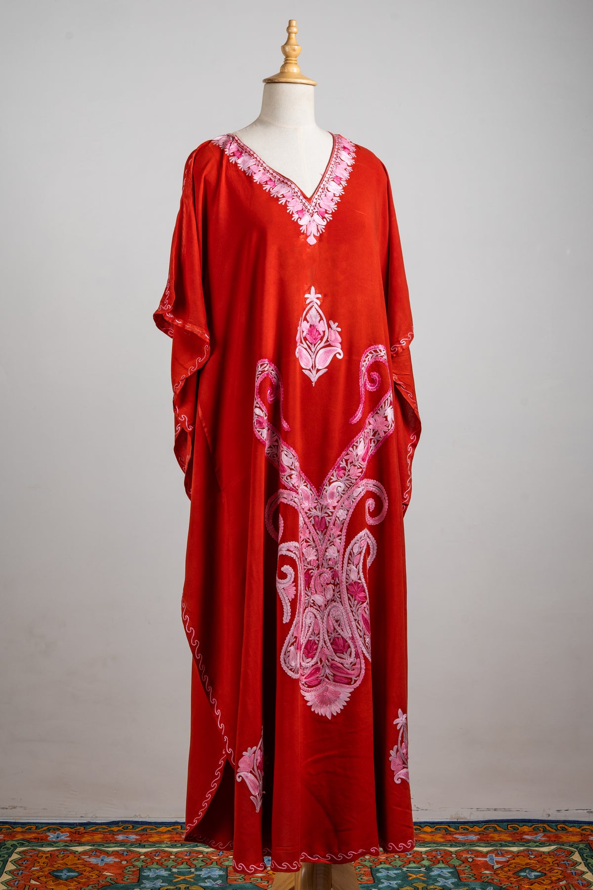 Full front view of the crimson kaftan with loose sleeves, a pink embroidered neckline and large central scroll design.