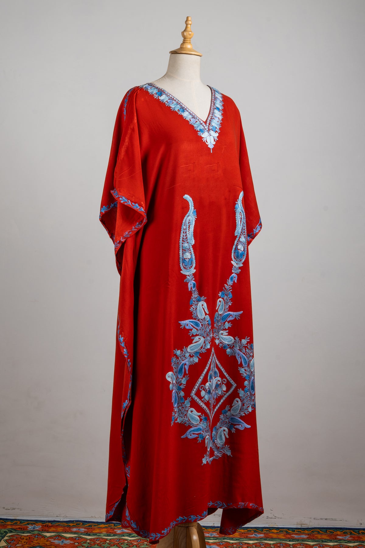 Full angled view of the red kaftan with a blue embroidered neckline, long paisley stems and a central diamond motif.