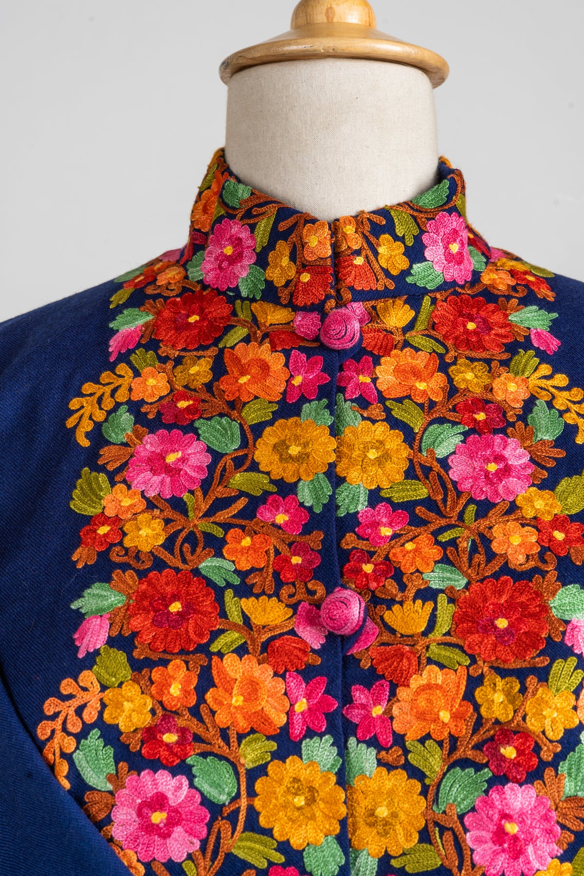 Detail of colourful Aari flowers and green leaves framing pink covered buttons on the navy jacket’s front