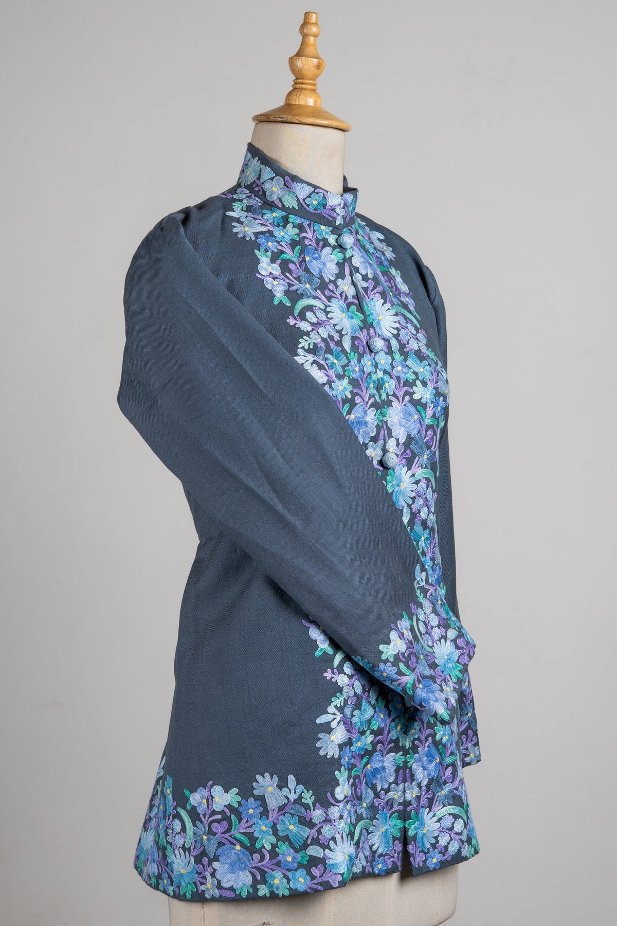 Angled view of the steel-grey jacket with one sleeve crossed, floral embroidery down the front and a wide decorated hem.