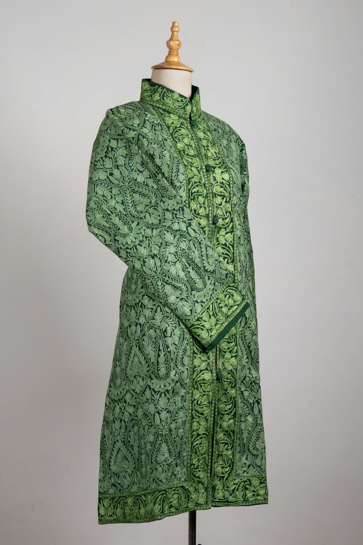 Angled green long jacket on a mannequin with dense tonal Aari embroidery, a stand collar and one sleeve crossed