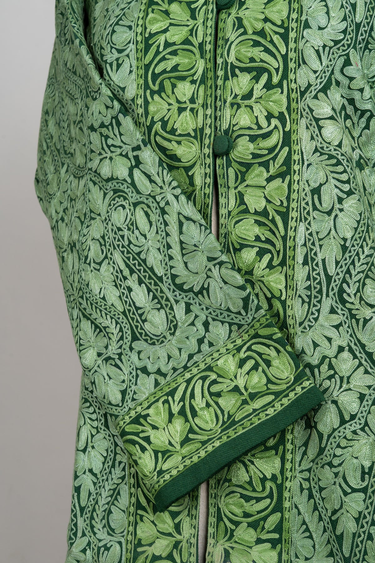 Close-up of a green jacket's crossed sleeve and buttoned front, with pale Aari paisleys and brighter leafy border embroidery