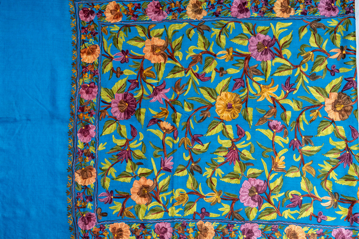 Flat end view of the peacock-blue stole, showing colourful Aari floral embroidery inside a border beside the plain end panel