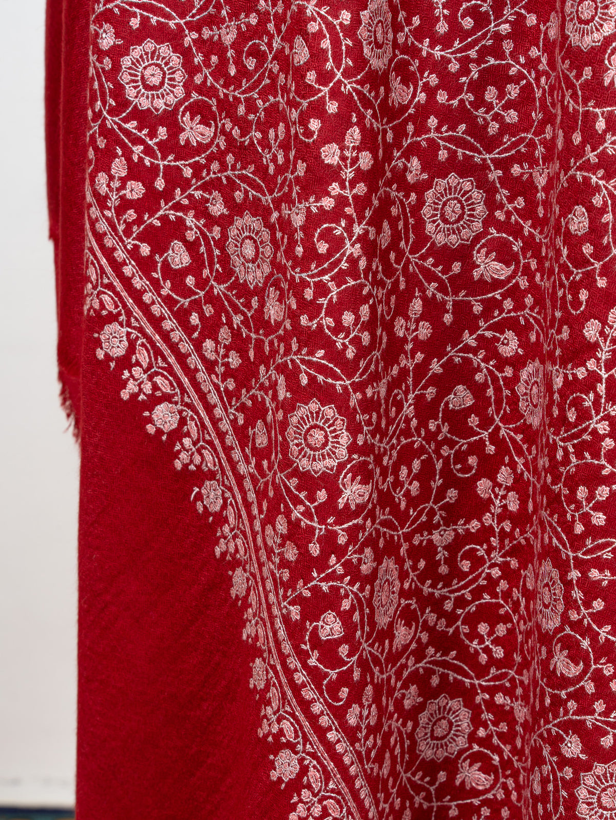 Hanging fold of a crimson shawl showing ivory Sozni floral vines and an ornamental border above a plain red end