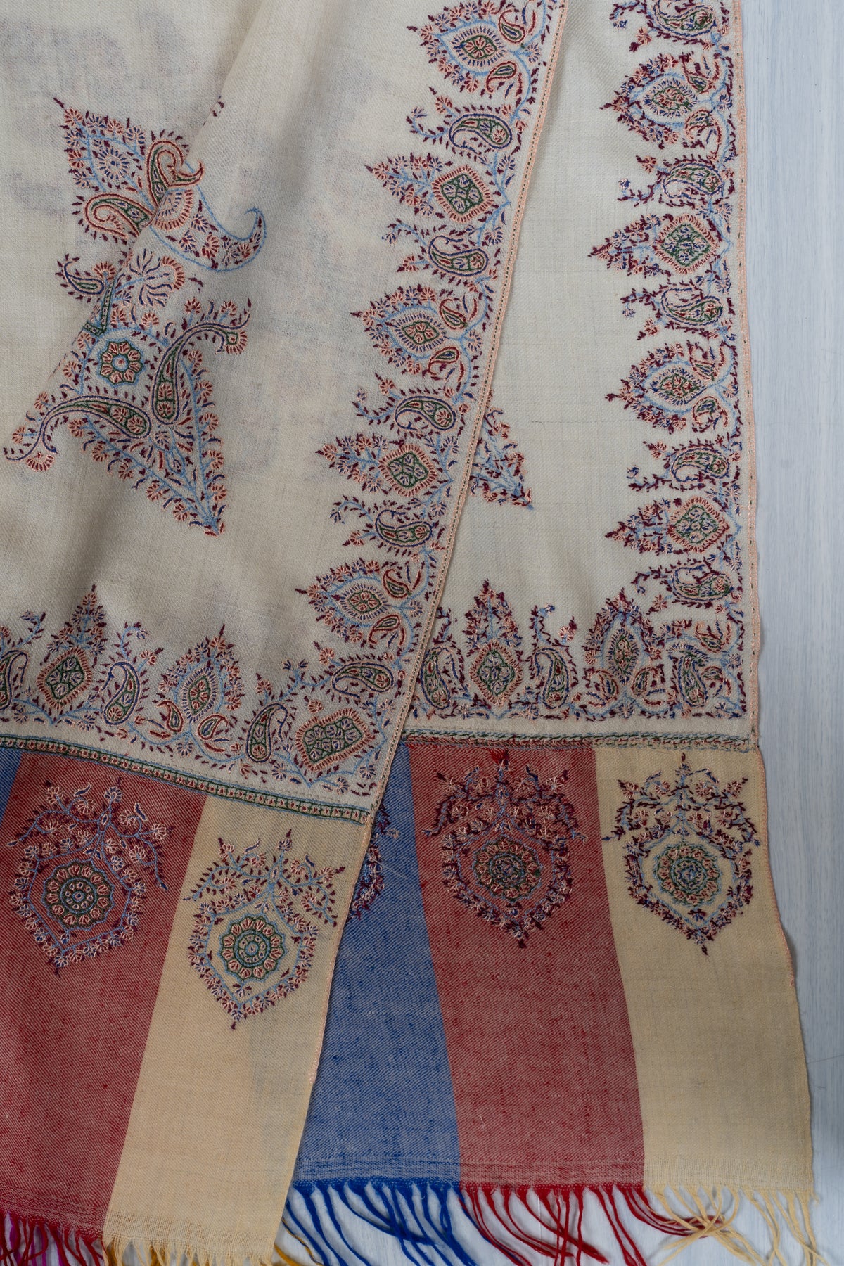 Overlapping ivory shawl corners showing ornate motifs, colourful end panels and blue and red fringe