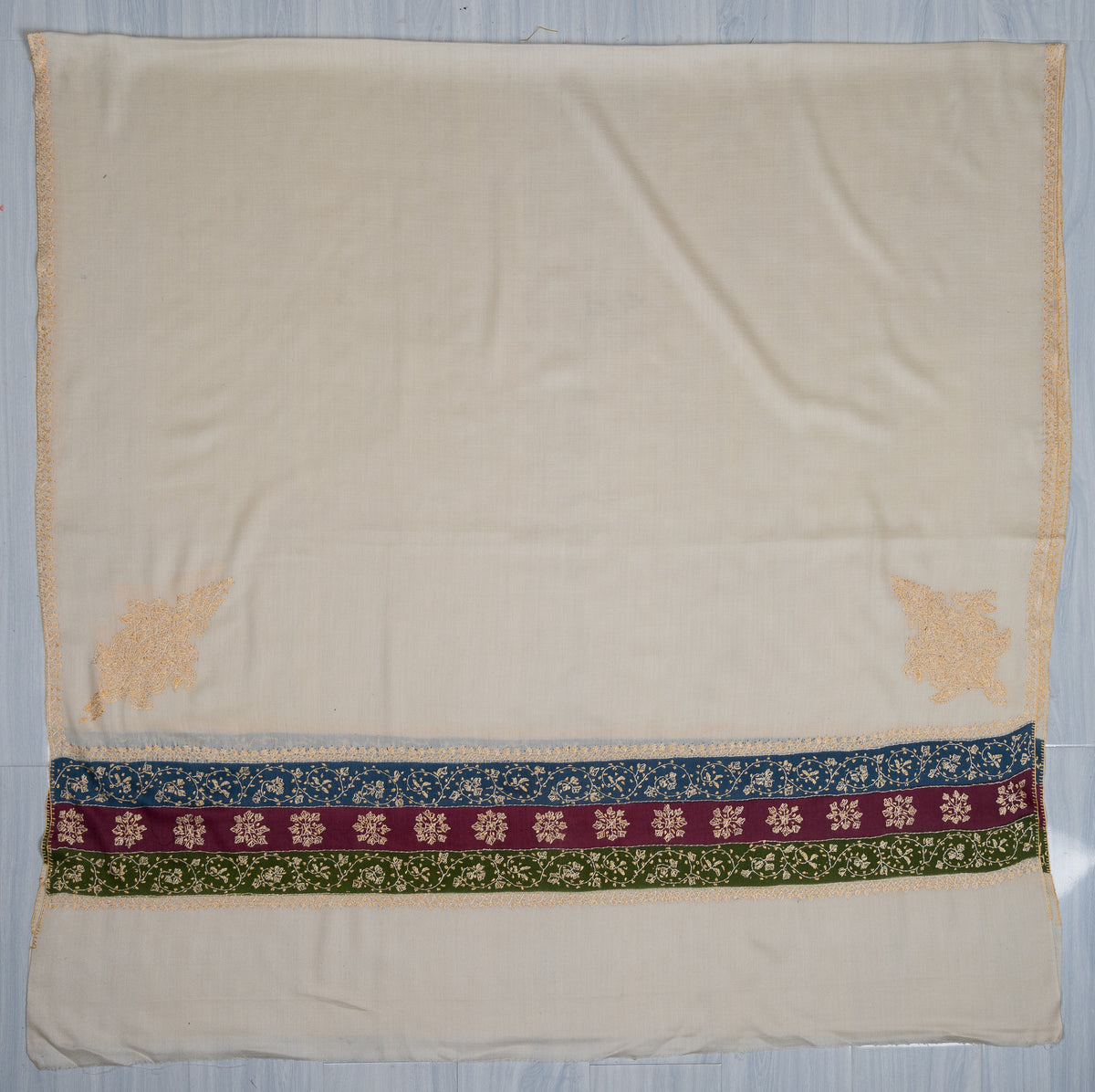 Ivory shawl laid flat with pale golden corner motifs and blue, maroon and green floral Sozni bands across the end