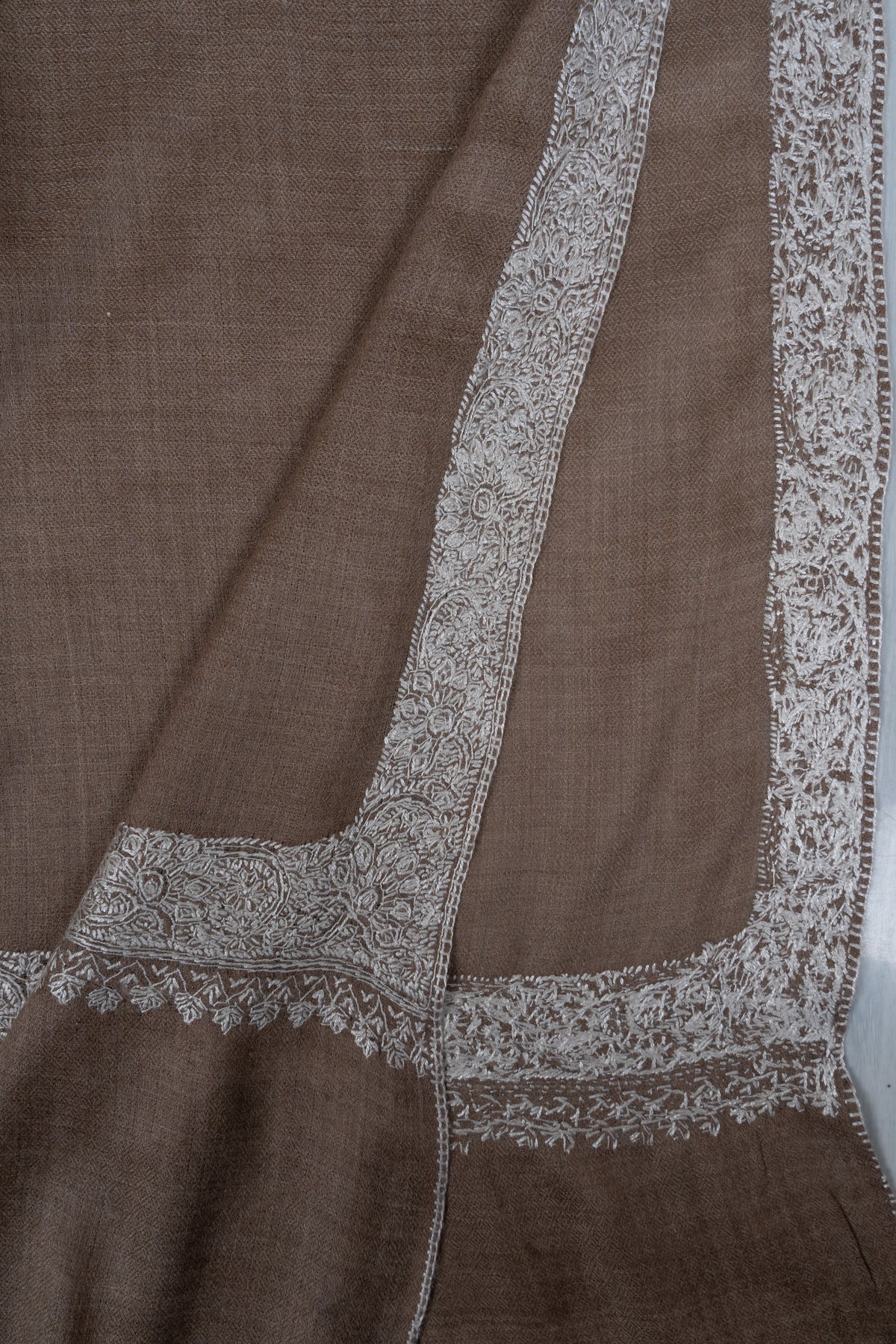 Folded corner of a walnut-brown Merino wool shawl showing the front and reverse of its silver embroidered border