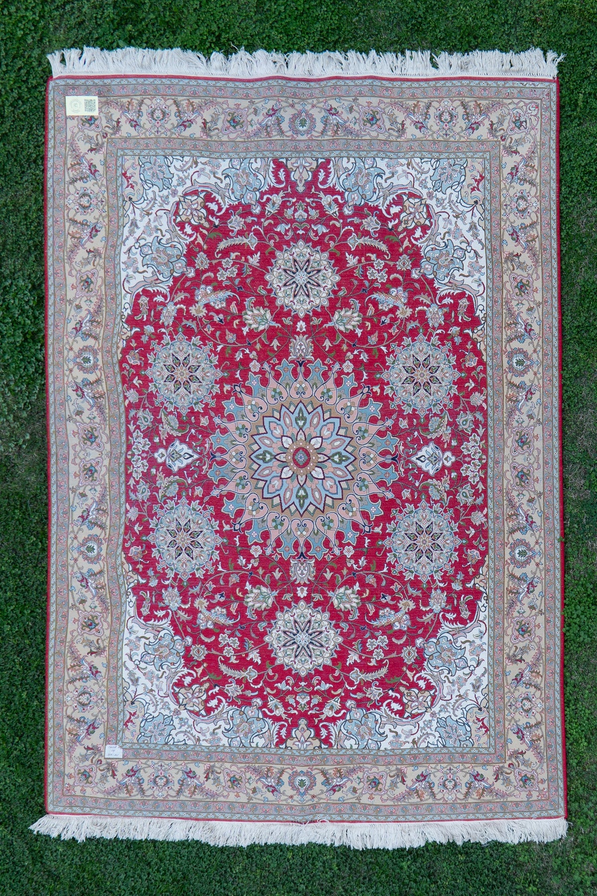 Reverse of a crimson floral carpet laid on grass, showing a central medallion, pale corner panels, beige borders, labels and white fringe