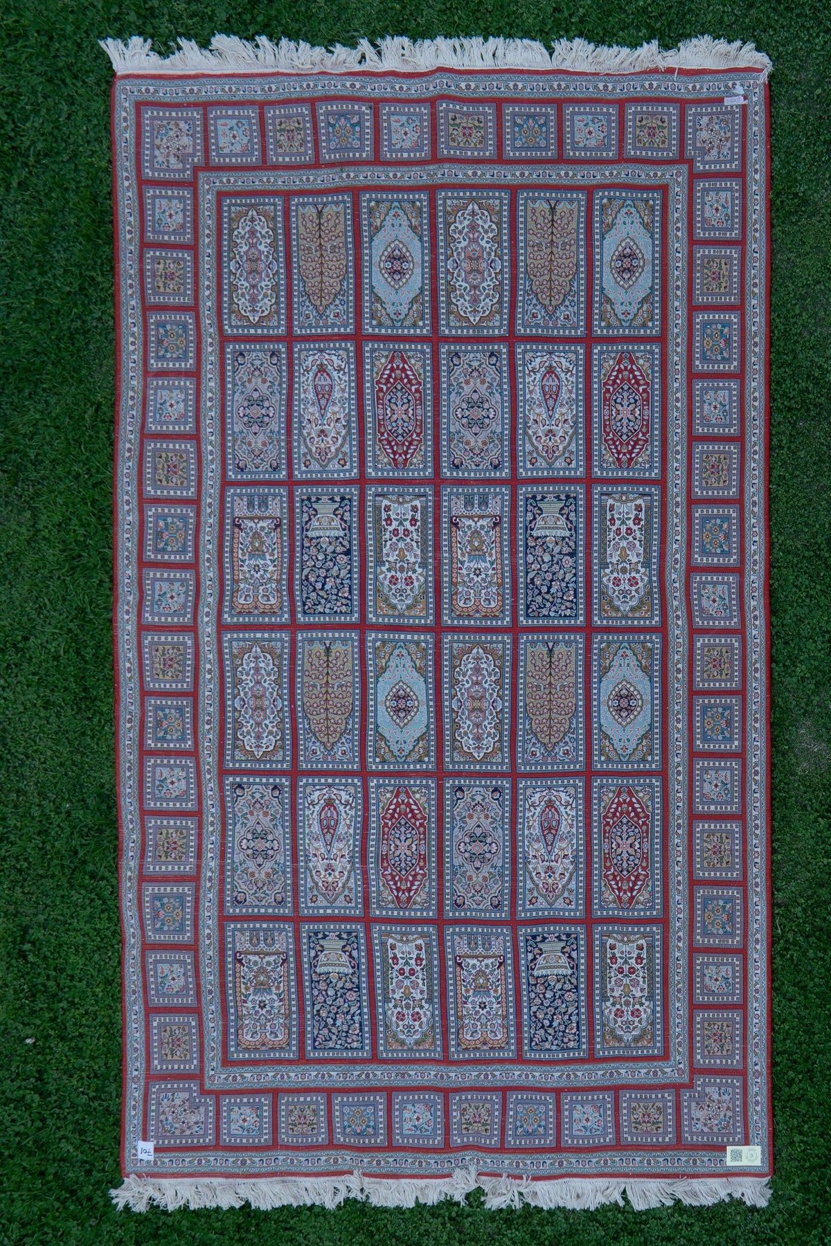 Reverse of the crimson-grid carpet laid flat on grass, showing rows of ornamental panels and white fringe