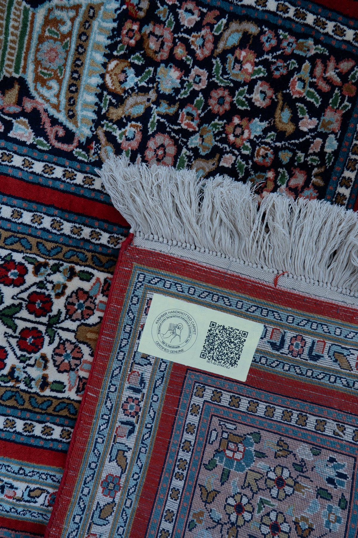 Folded carpet corner showing a QR label, white fringe and the woven reverse beside the floral face