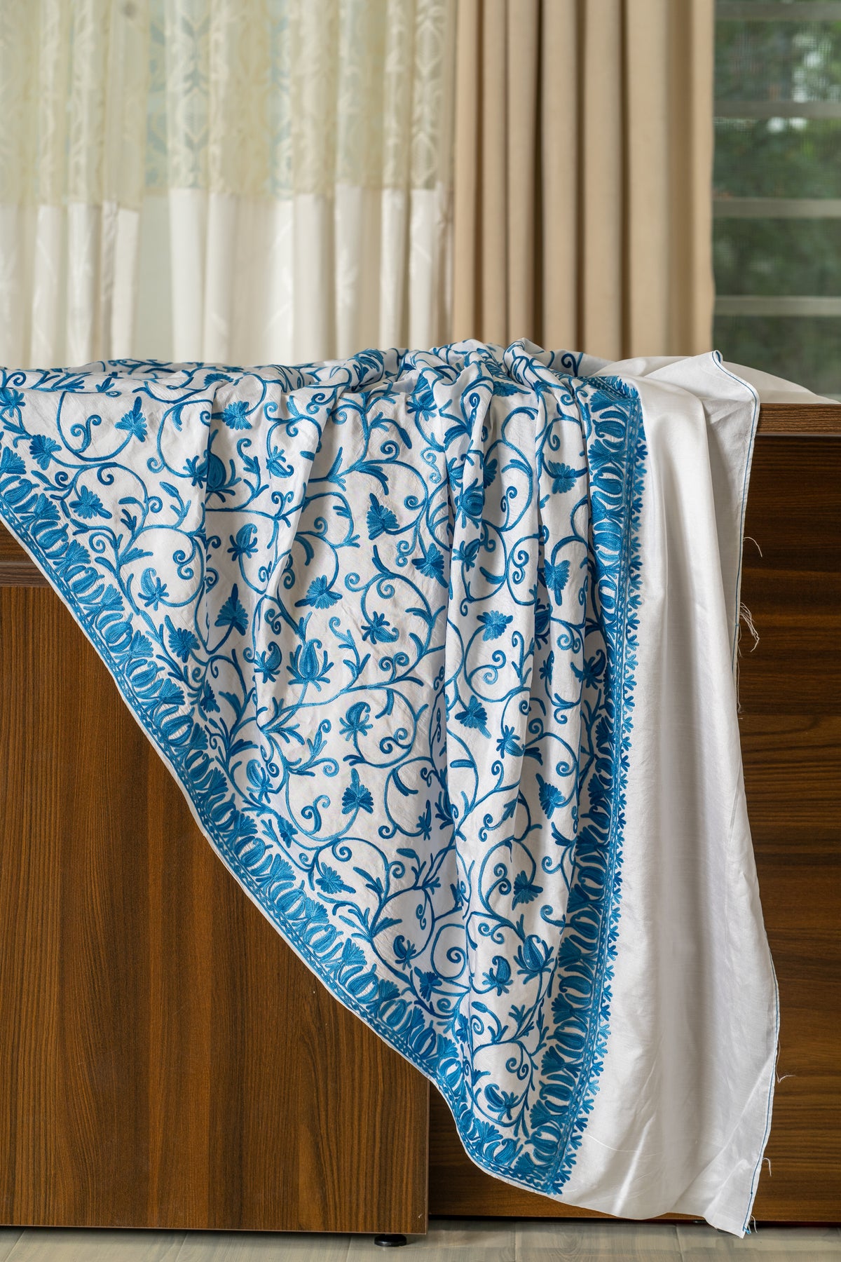Ivory saree draped across a wooden screen, with blue Aari floral vines, a leaf border and a plain ivory end