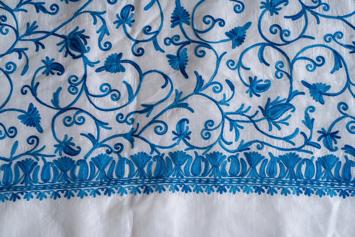 Blue Aari flowers and curling vines above an embroidered leaf border and plain ivory saree margin