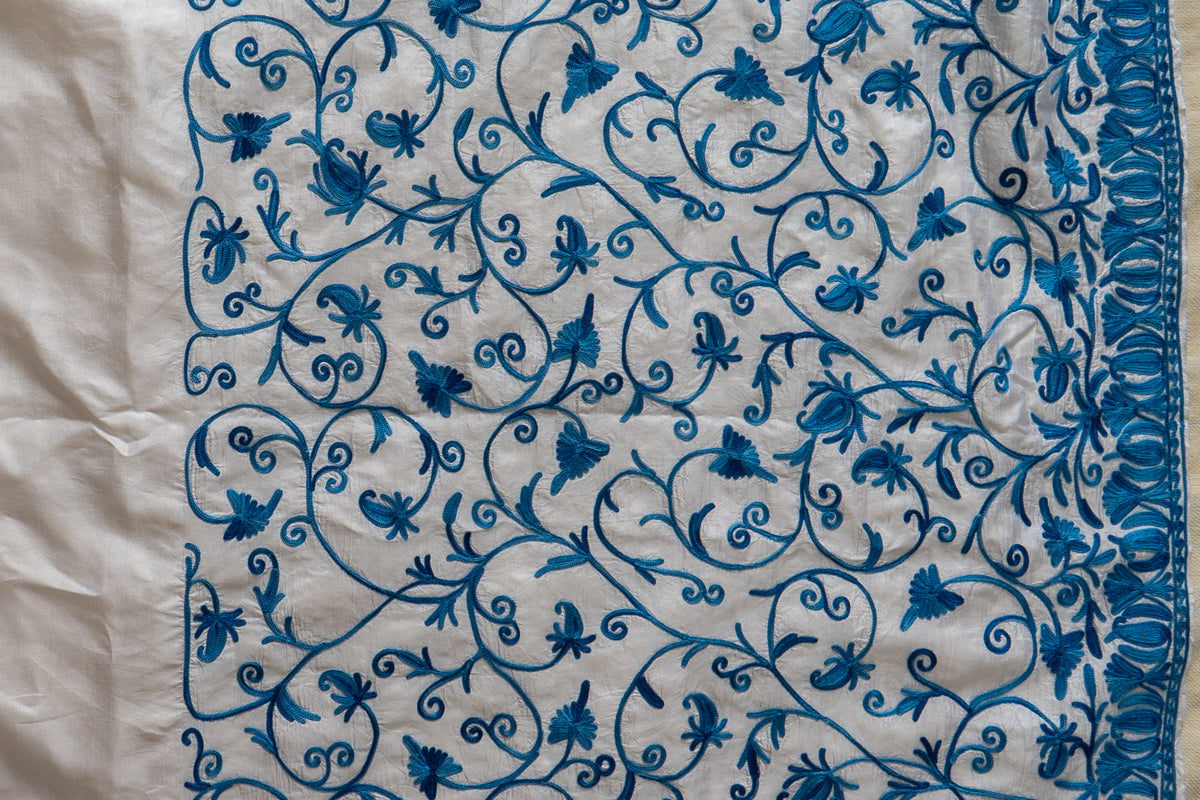 Detail of blue Aari floral vines beside a dense embroidered leaf border and plain ivory fabric
