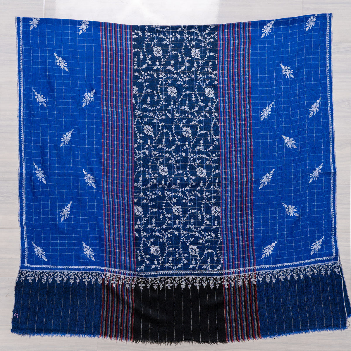 Royal-blue Pashmina shawl spread flat, with a dense white floral centre panel, checked sides, coloured stripes and a dark end