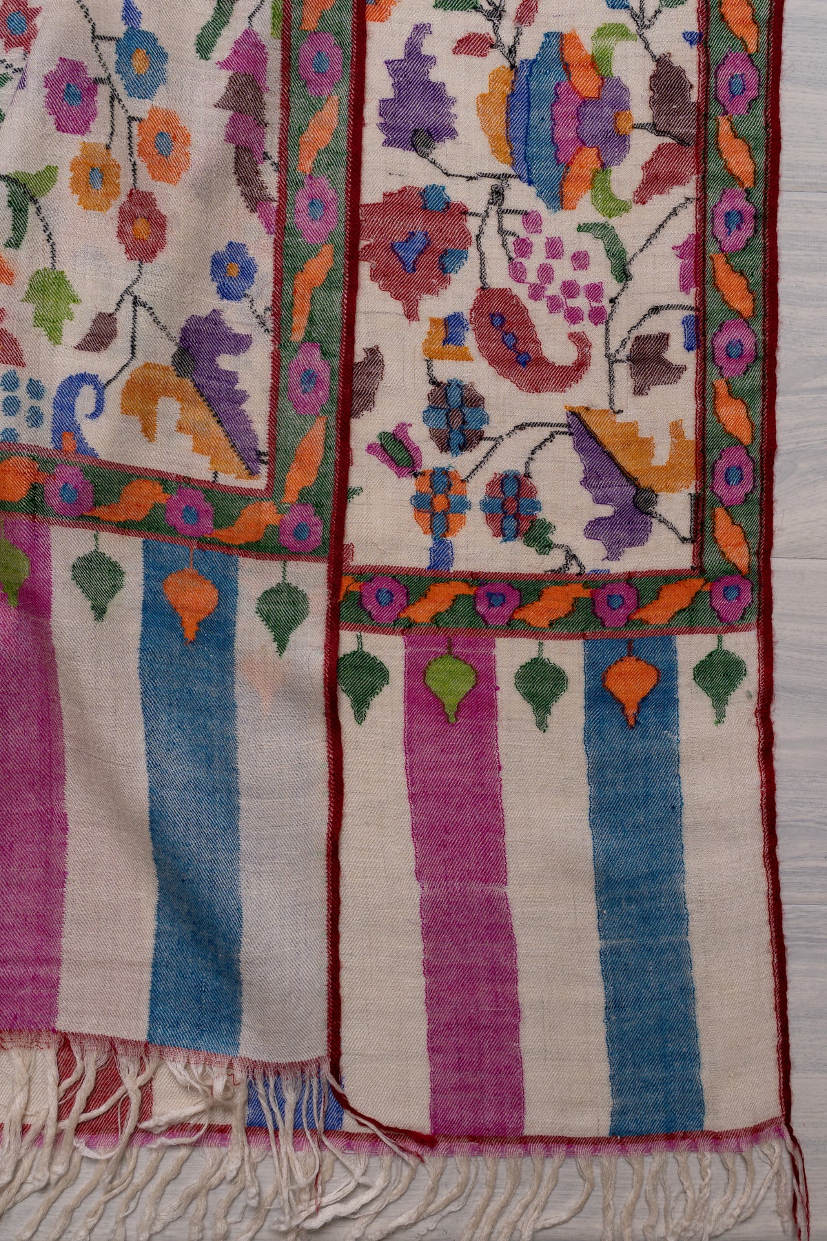 Overlapping ends of an ivory Pashmina shawl, showing colourful floral borders, pink and blue stripes and ivory fringe.