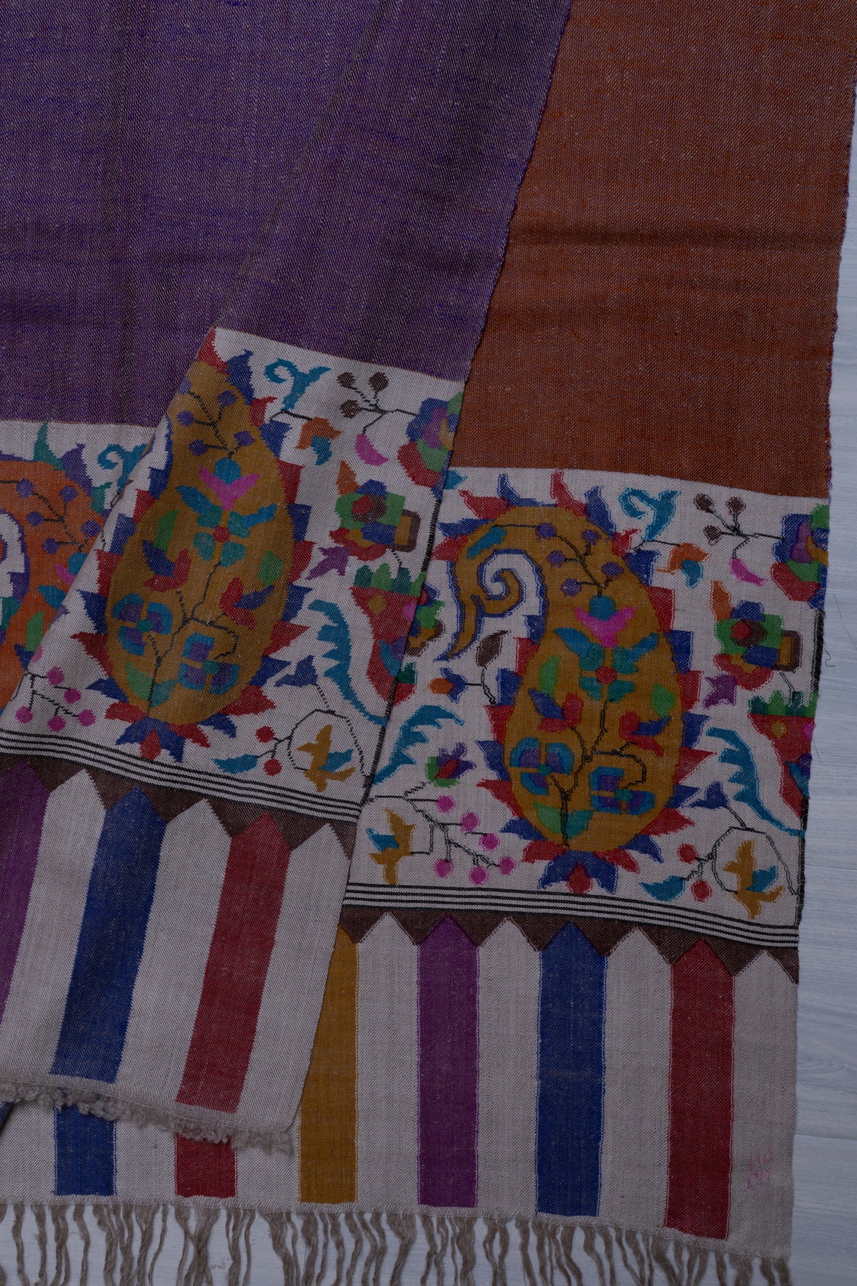Overlapping purple and rust-toned ends of a Pashmina shawl, with orange paisley panels, multicoloured stripes and ivory fringe.