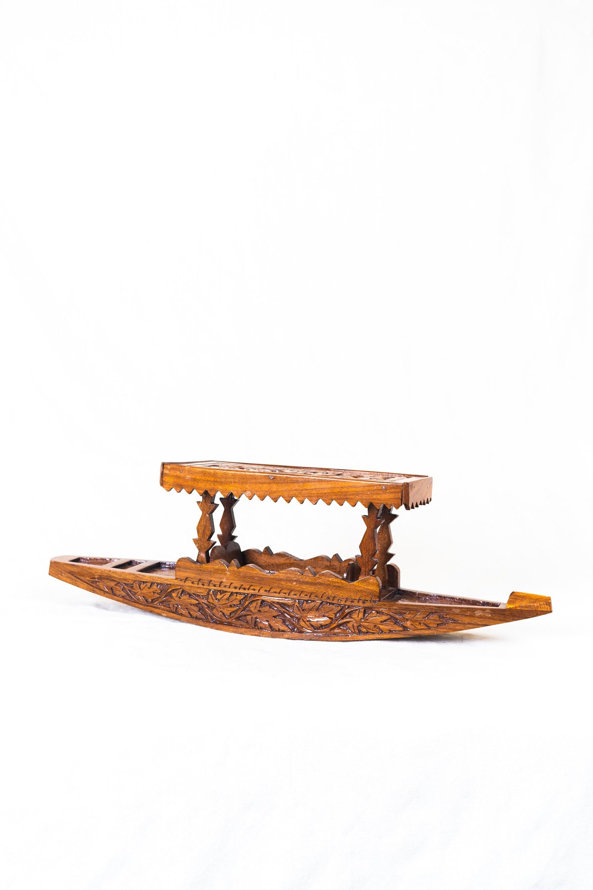 Angled side view of a polished walnut wood shikara model with leaf carvings along the hull and scalloped canopy trim