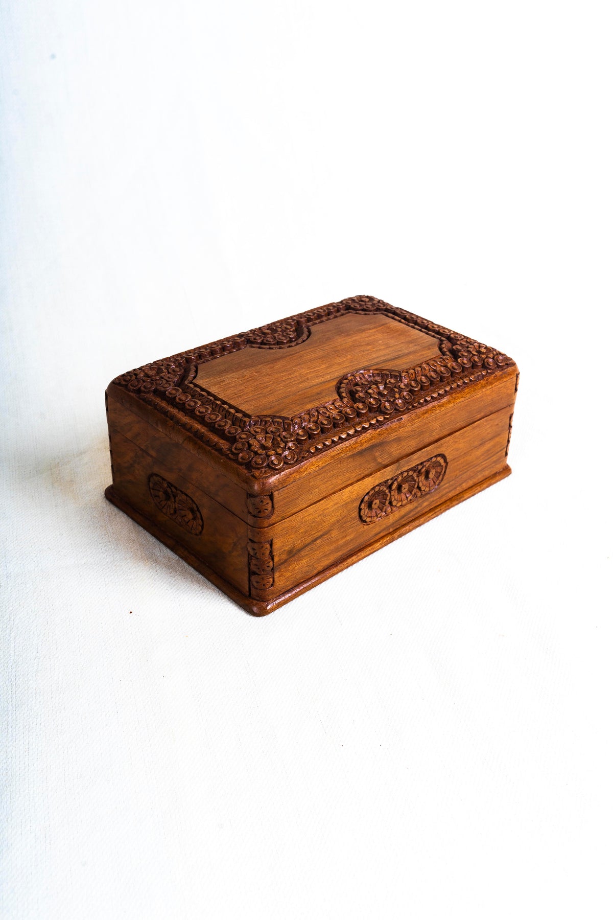 Opposite angled view of the walnut box, showing its plain wood-grain lid centre framed by carved flowers.